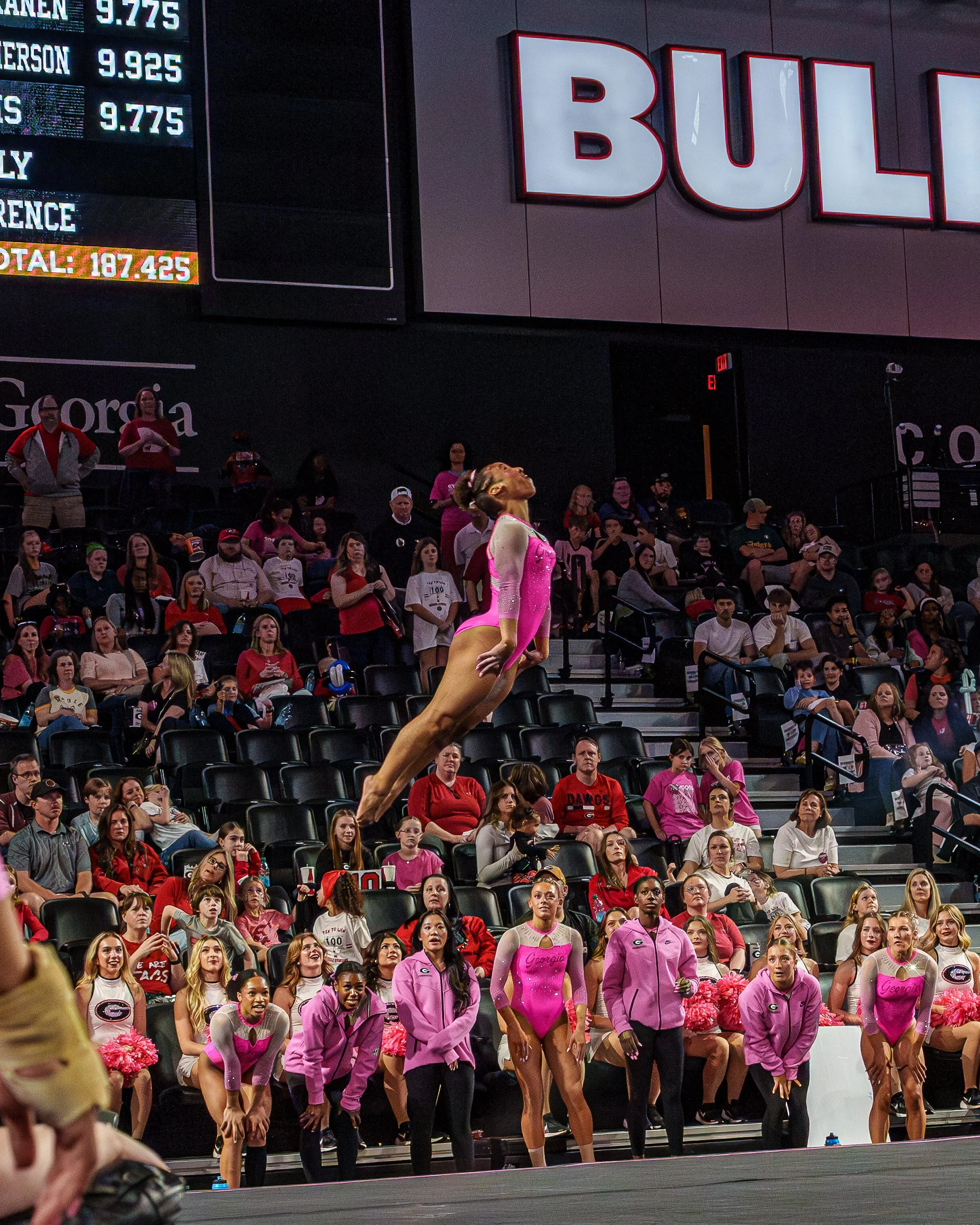 An artistic gymnast wearing a pink leotard performs a leap in front of an audience at a gymnastics competition. The audience is watching attentively, some standing, some seated, in an indoor arena. Cheerleaders in pink and white uniforms are seated in the front row.