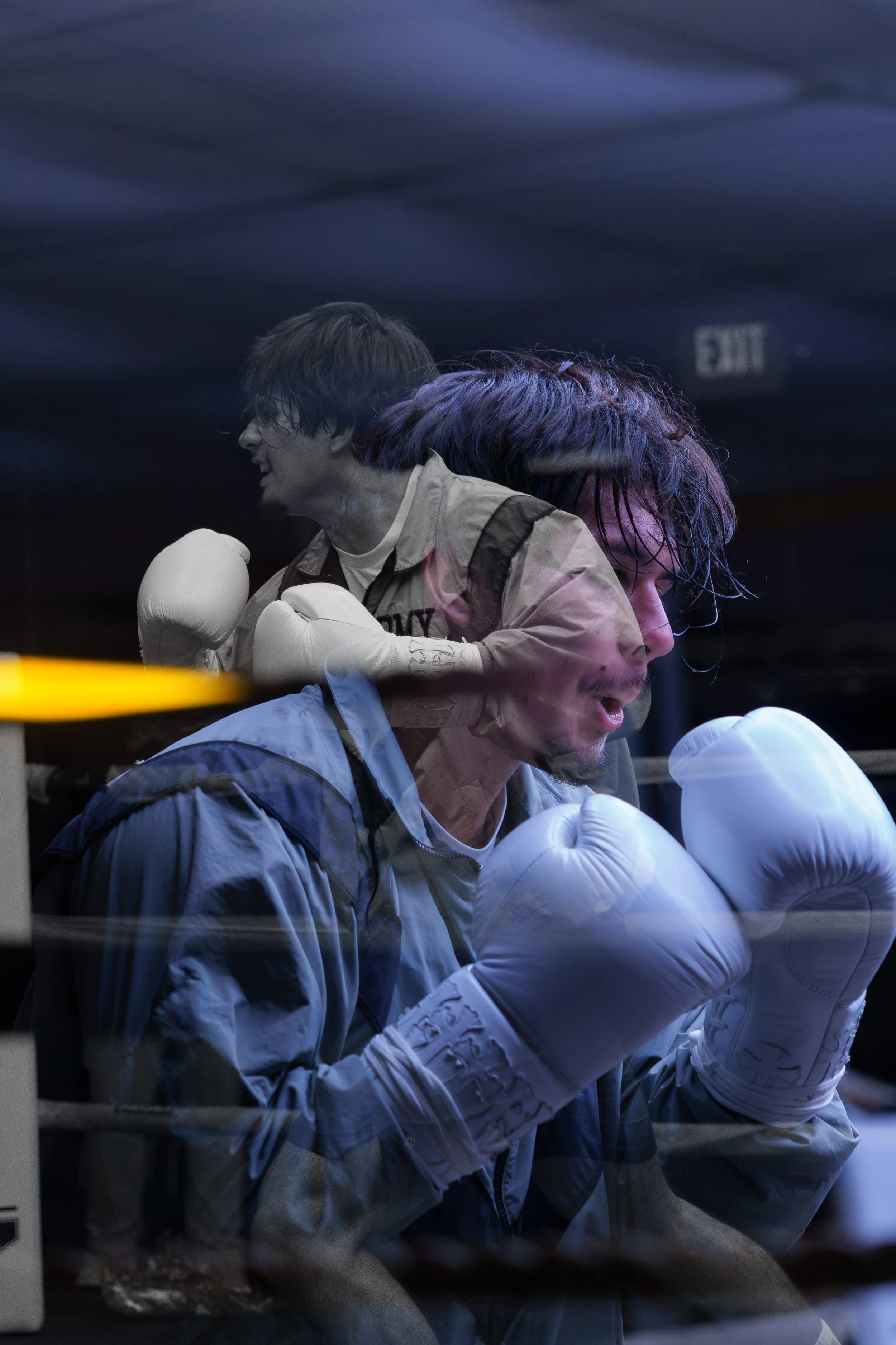 A young male boxer with wet hair and wearing white boxing gloves, deep in concentration inside a boxing ring, with a reflection of a boxer in the background, all captured through a window with reflections.