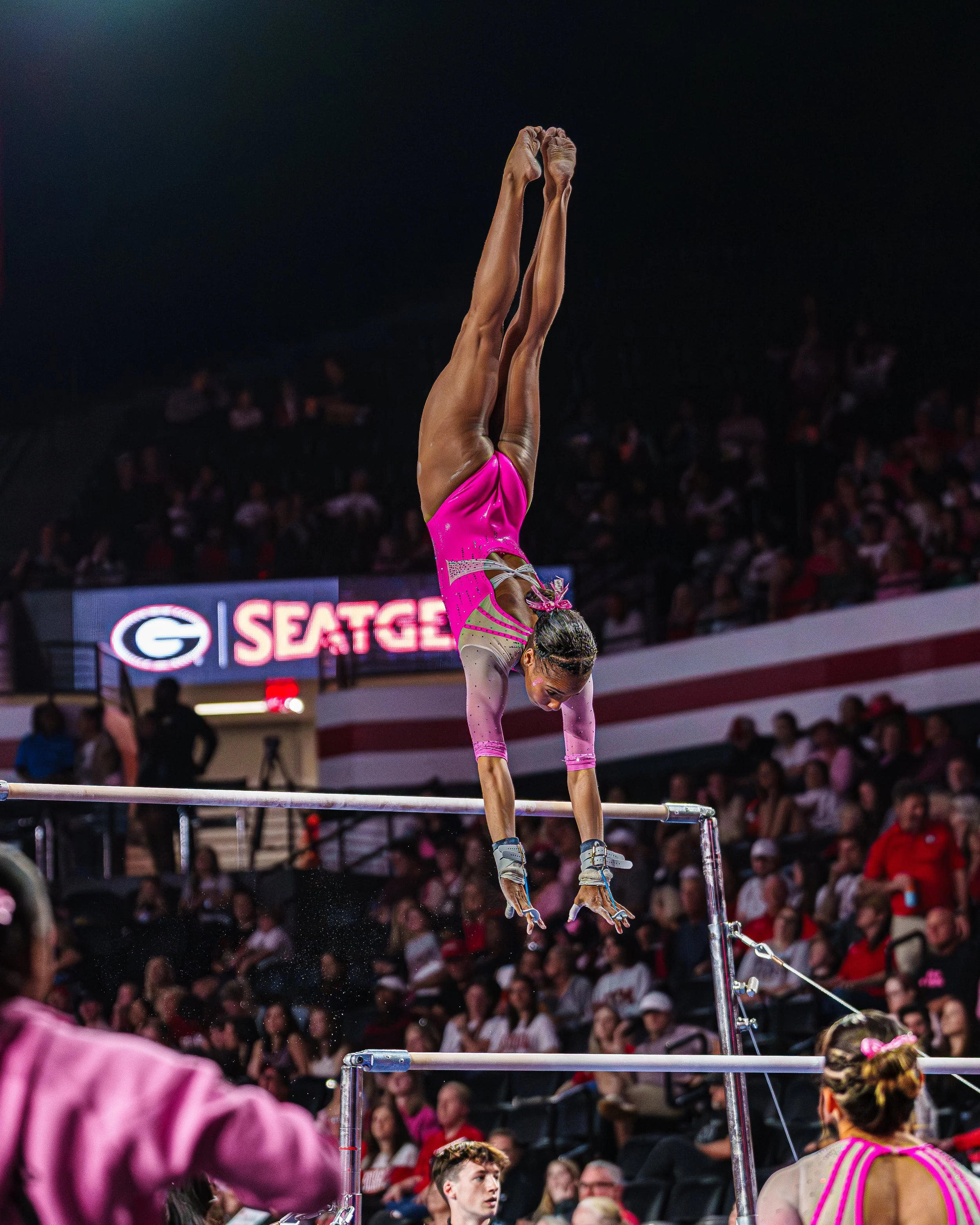 An female gymnast in a pink and green leotard performs a handstand on the uneven bars during a gymnastics competition, with a large crowd watching in the background.