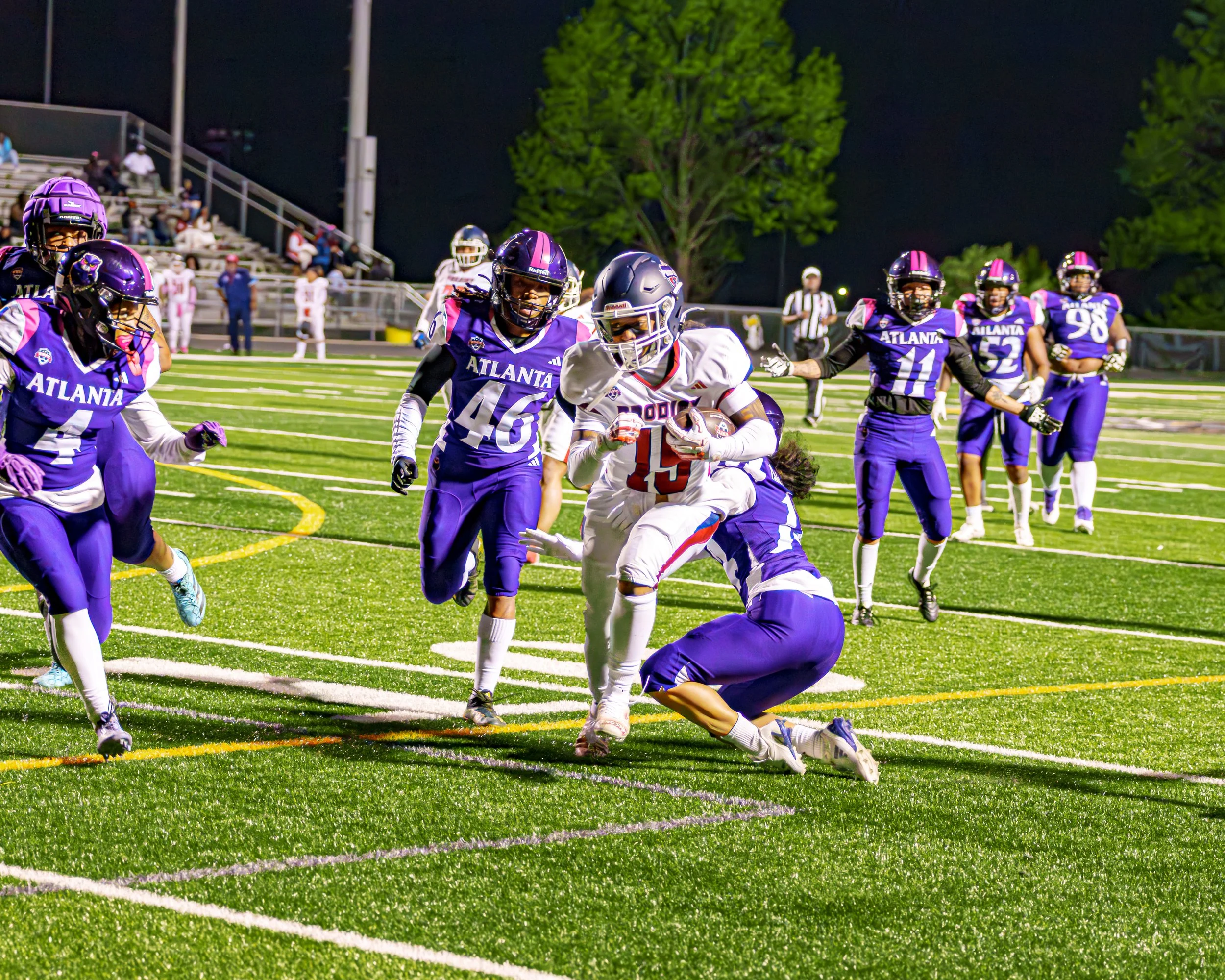 American football game at night, players on field wearing purple and white uniforms, one player running with the ball.