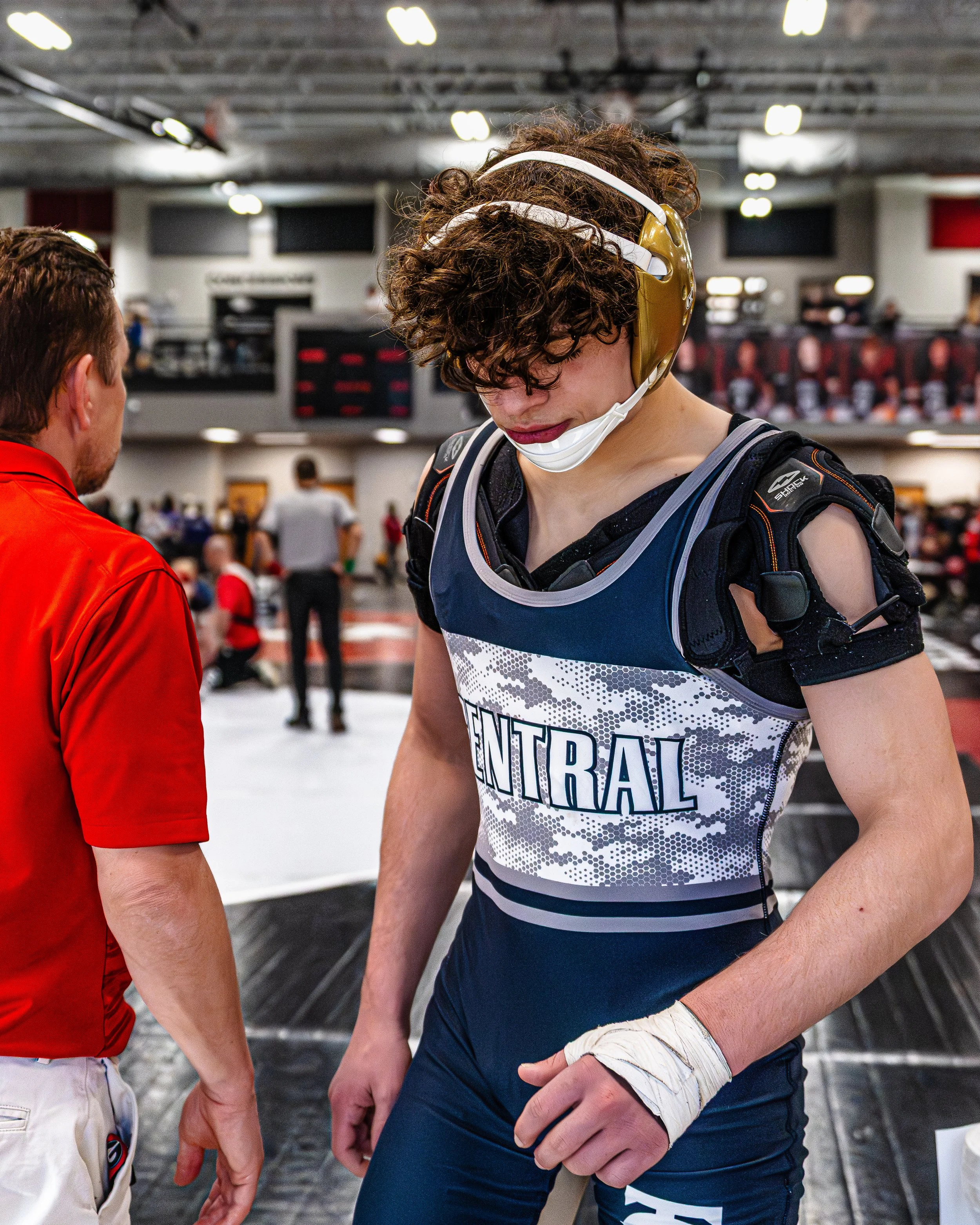 Young male wrestler wearing a blue and white singlet with 'CENTRAL' printed on it, a protective headgear, and a wristband, standing in an indoor sports arena.