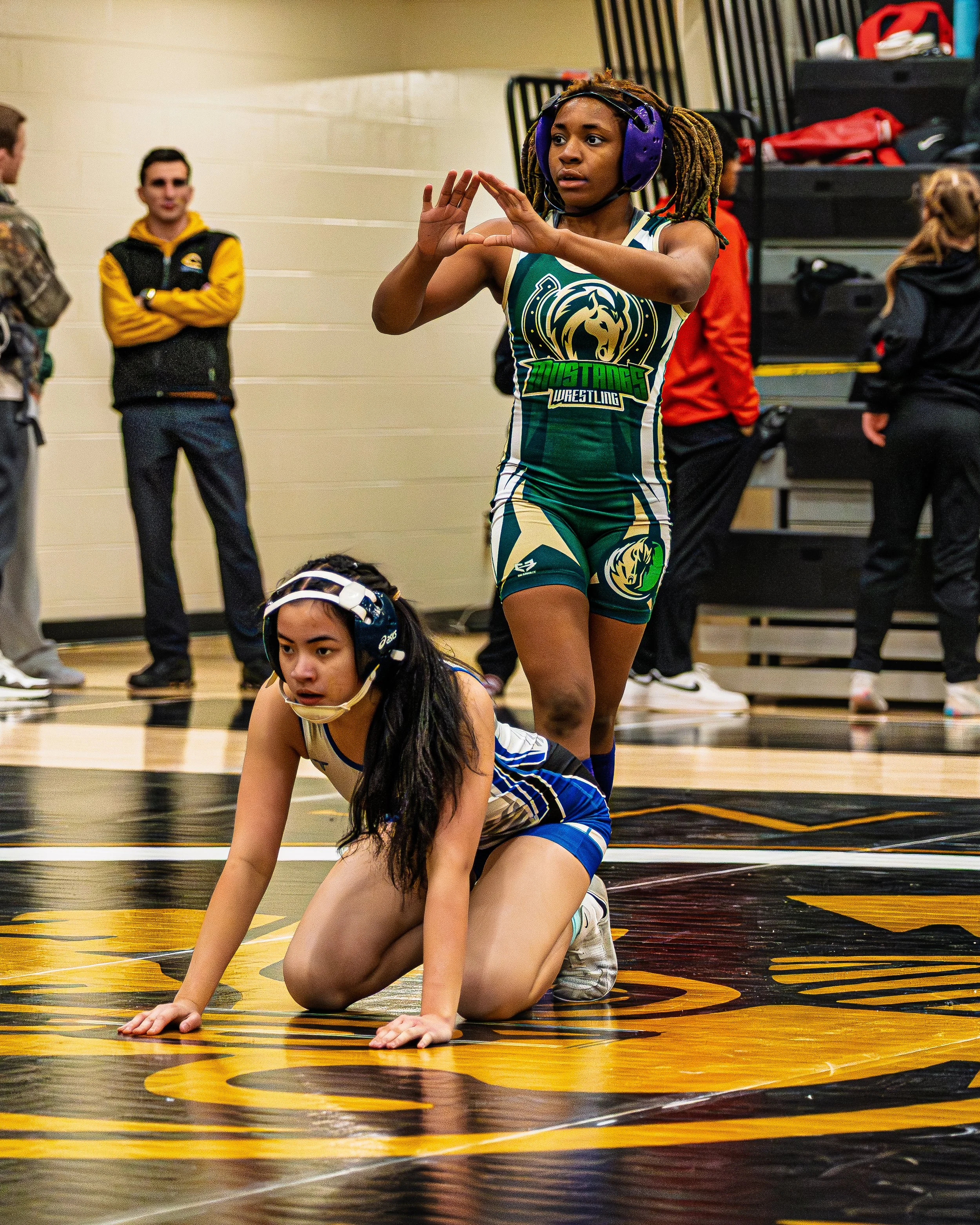 Wrestling match with a female wrestler in green and gold uniform standing with arms raised and another female wrestler in blue and white uniform on all fours on the wrestling mat, with spectators in the background.