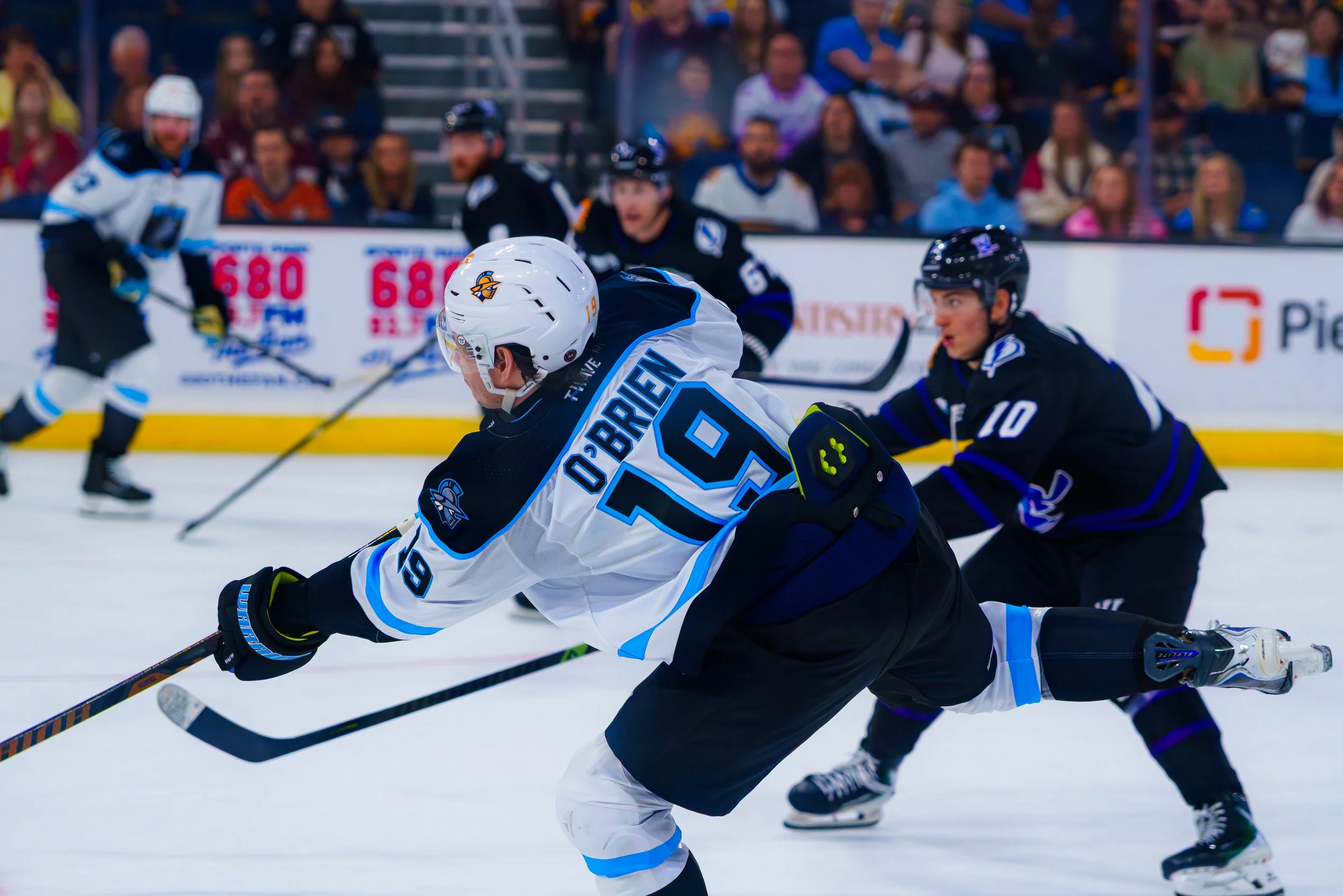 Hockey players in action during a game, with one player leaning forward reaching for the puck while others skate behind him.