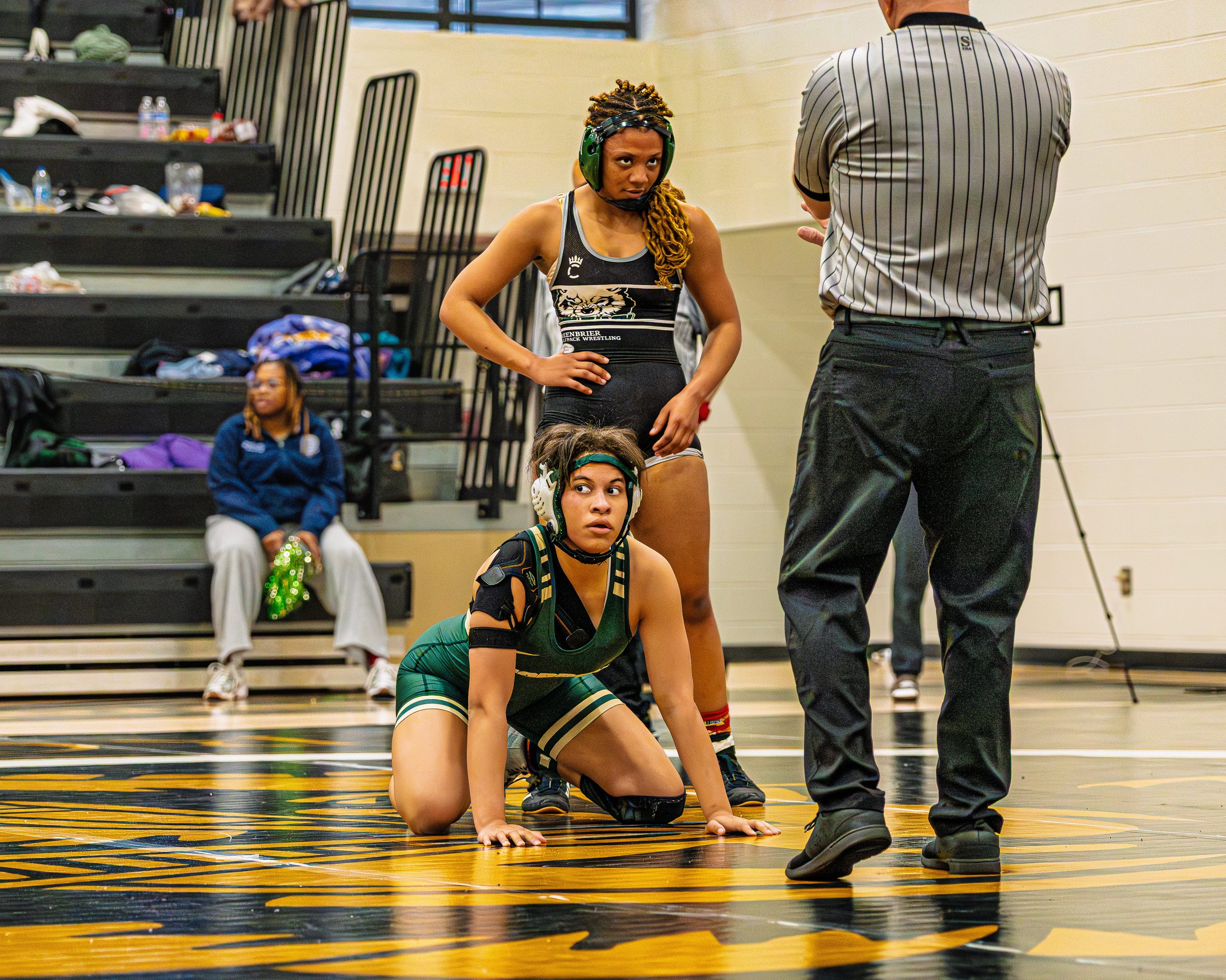 Two female wrestlers in black and green uniforms kneeling and standing on a wrestling mat, looking at a referee. A girl in a blue jacket sits on a bench in the background in a gymnasium.