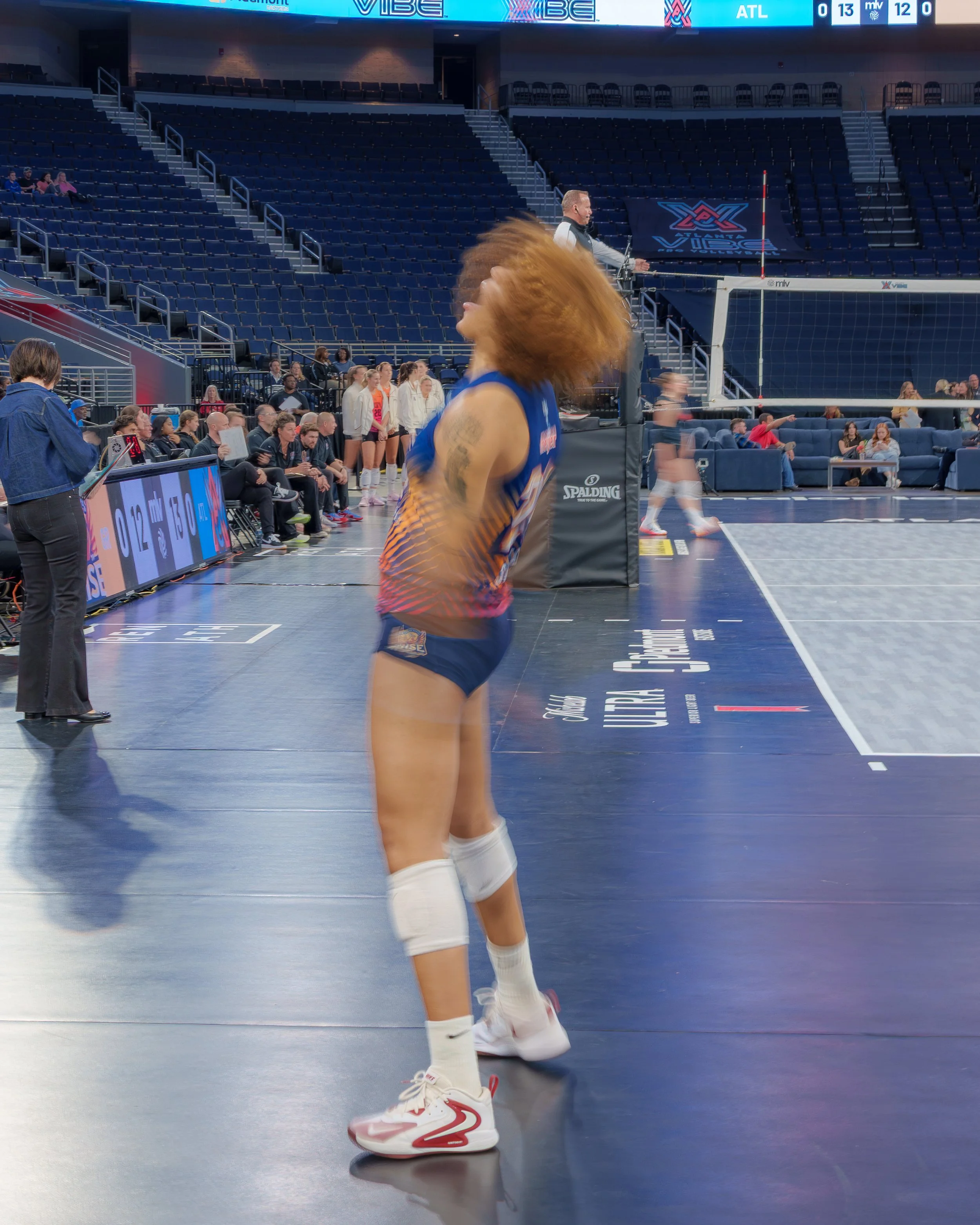 A female volleyball player with red hair, tattoos on her arm, wearing a blue and orange uniform, white knee pads, and white sneakers, standing on an indoor volleyball court with a volleyball net in the background, and people sitting and standing in the stands.