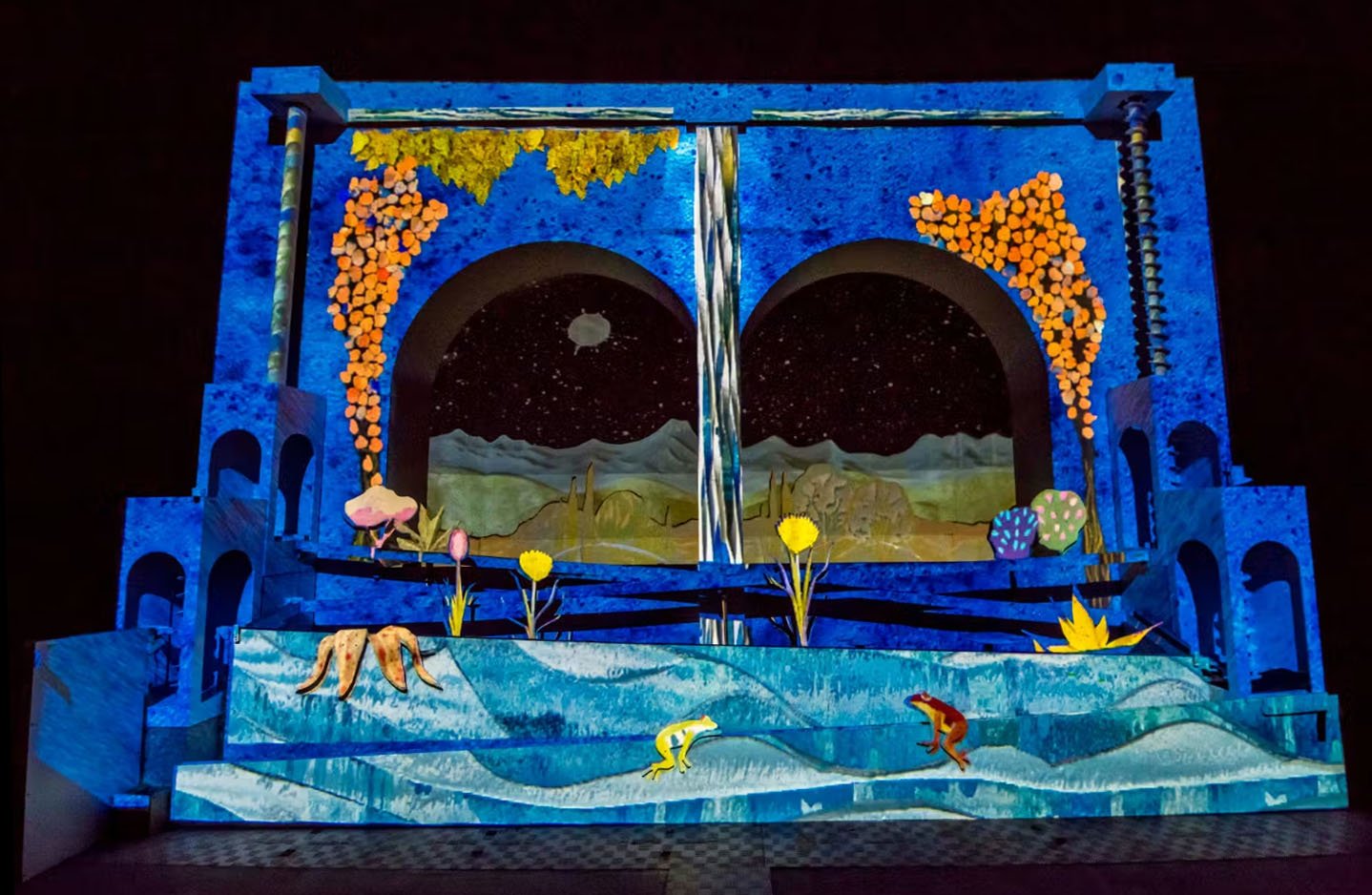 Colorful stage set with a blue structure resembling a castle or theater, featuring arches and columns, decorated with flowers and trees, a painted mountainous landscape background under a starry night sky, with frogs and a turtle on a water surface in the foreground.