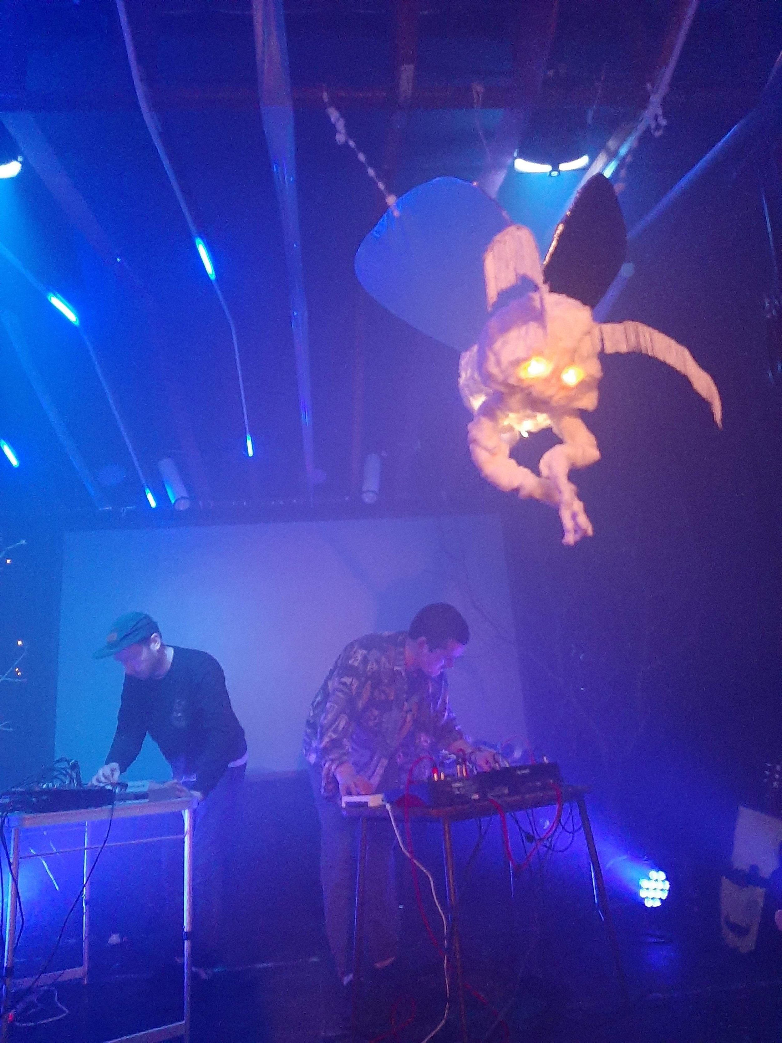 Two people are performing live music on stage with electronic equipment. Above them, a large illuminated creature with glowing eyes, wings, and a tail is suspended from the ceiling.