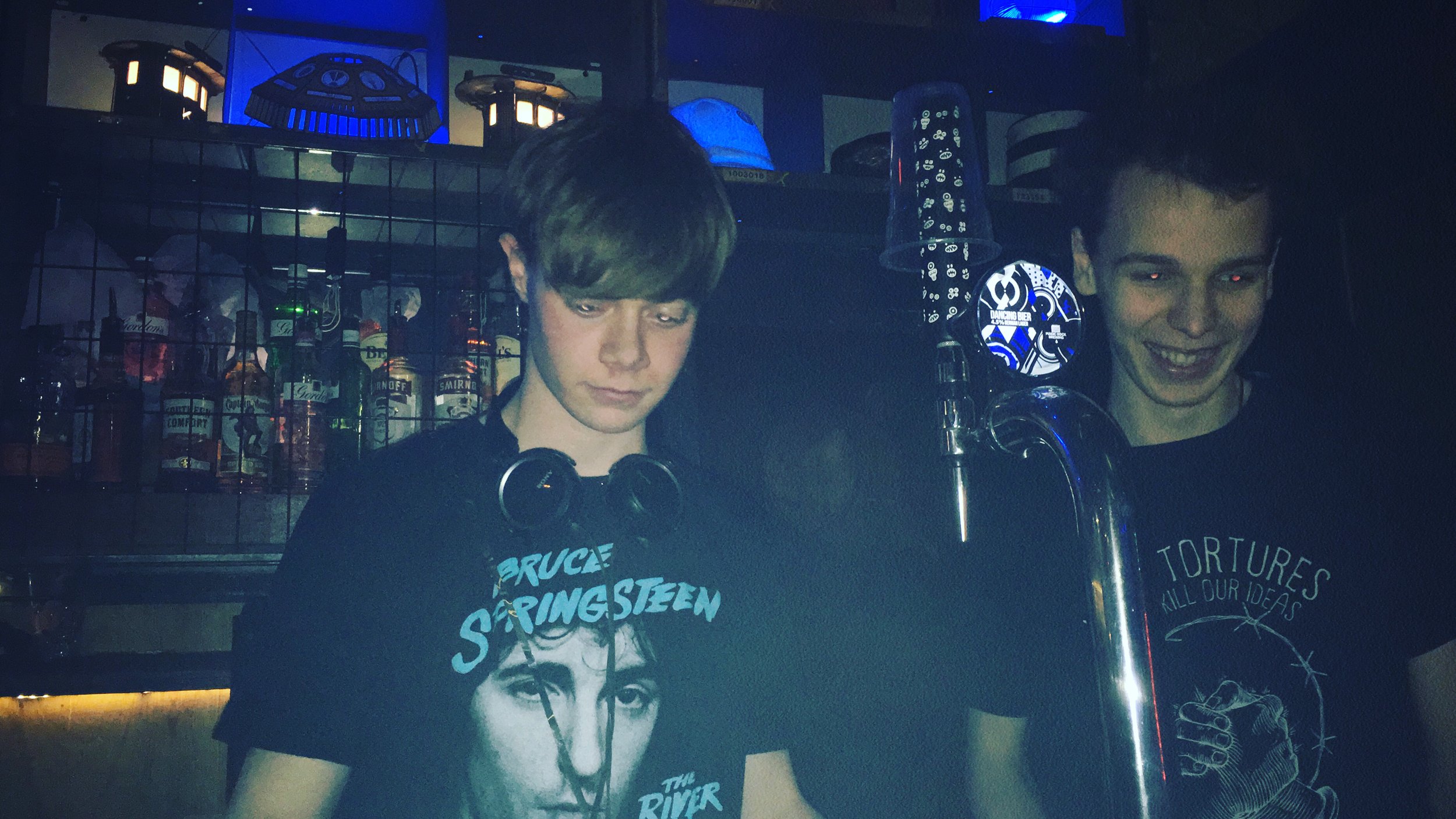 Two young men with short hair, one with dark and the other with red eyes, standing behind a bar or DJ booth with liquor bottles in the background. The man on the left is wearing a Bruce Springsteen T-shirt and headphones around his neck, looking down