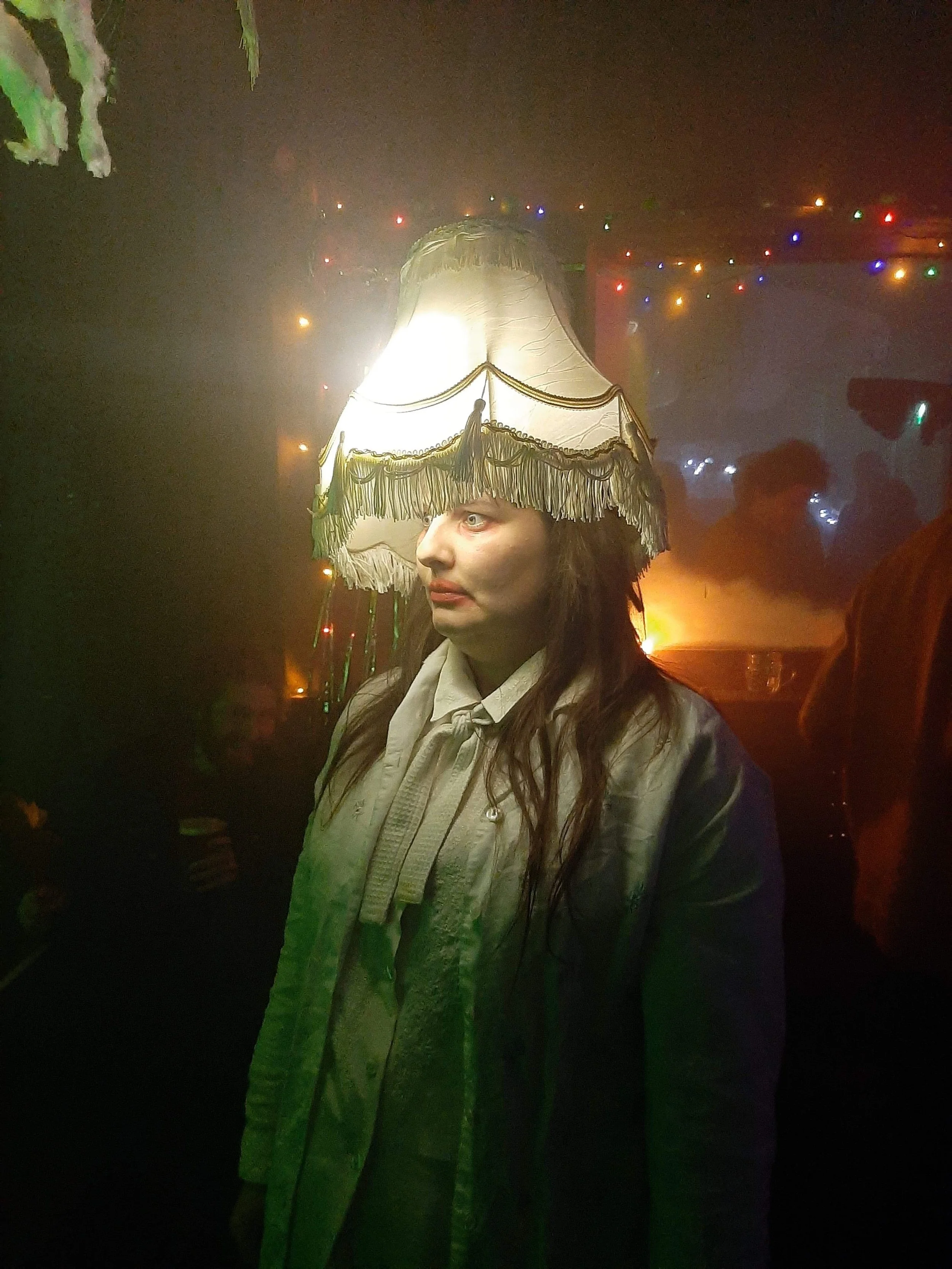 A woman with pale skin and long dark hair wearing a vintage style white shirt and a coat, illuminated by a large, old-fashioned lampshade on her head, in a dimly lit room with colorful string lights and smoky atmosphere.