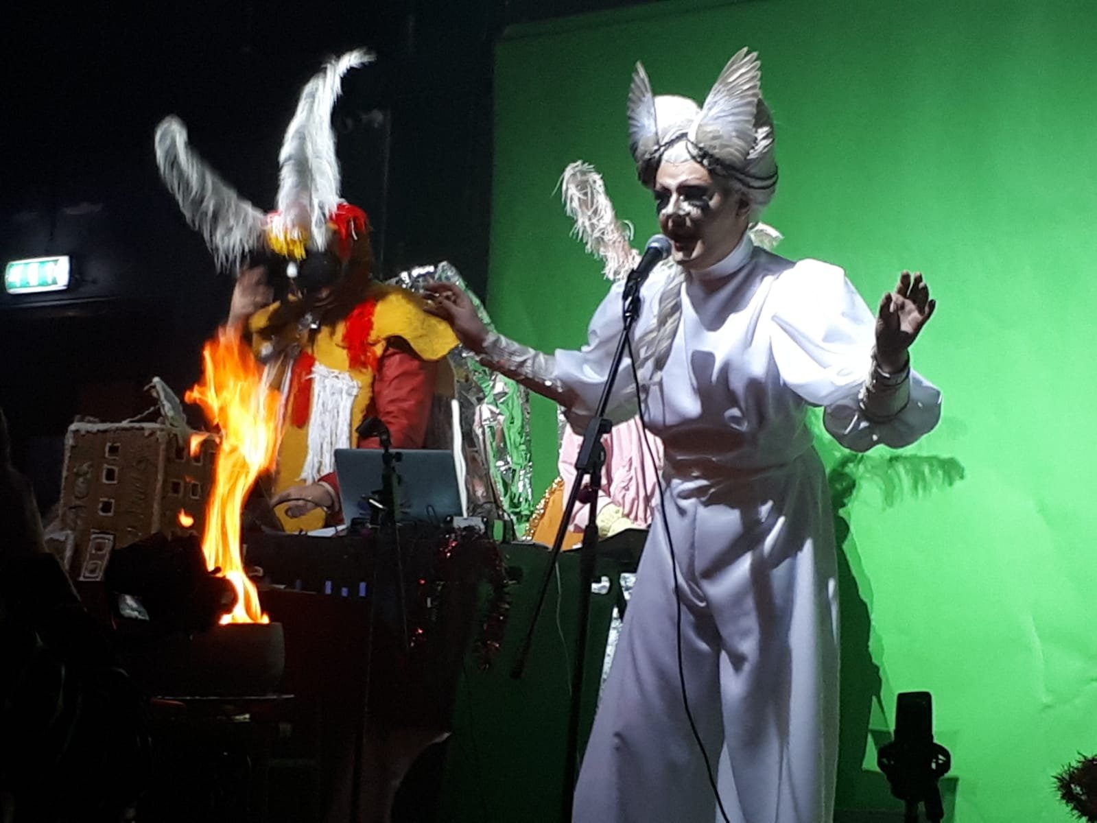 Two performers dressed in theatrical costumes on stage, one with a white dress and elaborate headpiece singing into a microphone, the other in colorful attire with a feathered mask, standing near a flame and a set piece with a green screen background