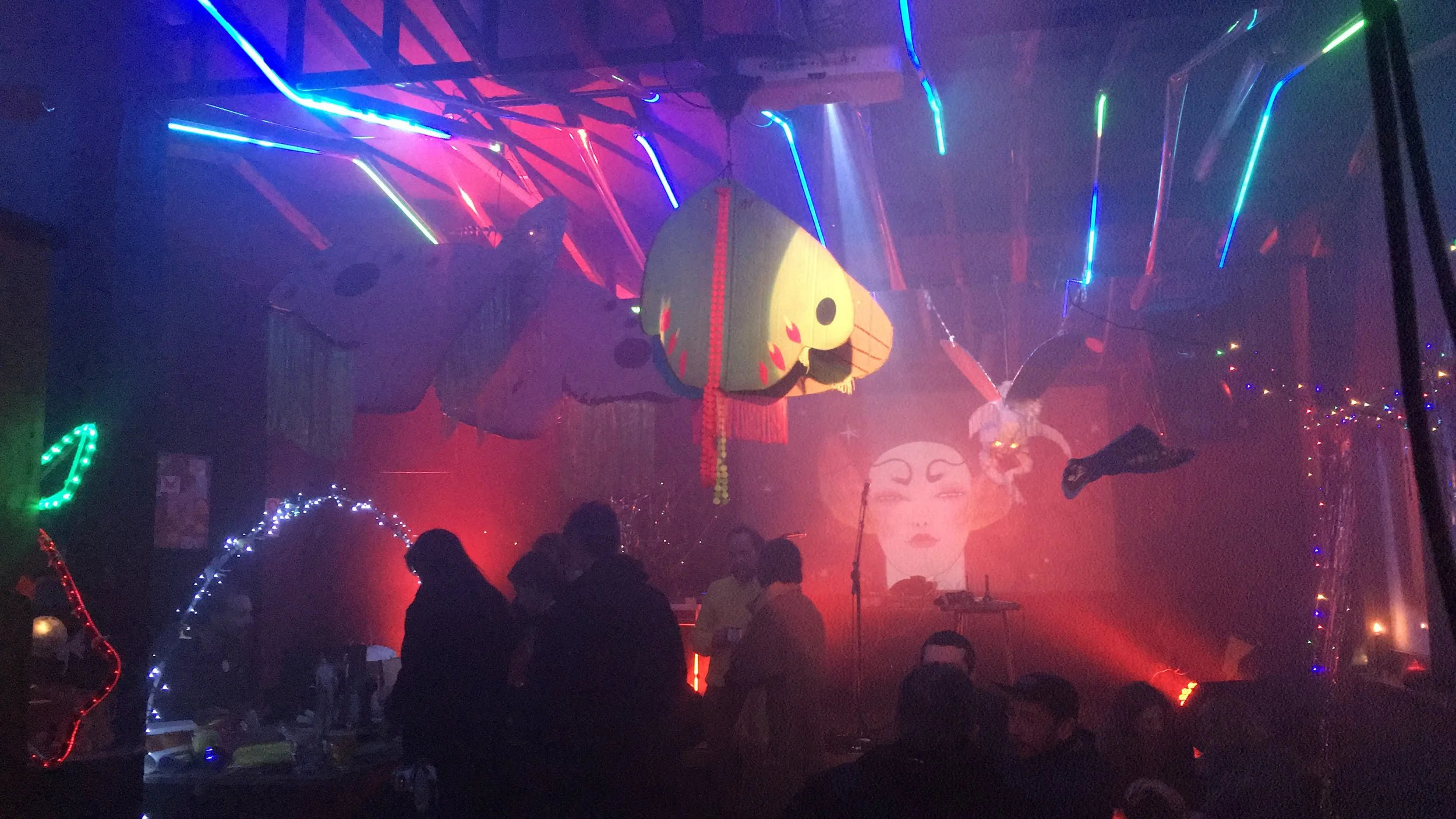 A dark, colorful venue with neon lights, large fruit-shaped lanterns, and a projection of a woman's face on the back wall. Silhouettes of people are visible enjoying the event.