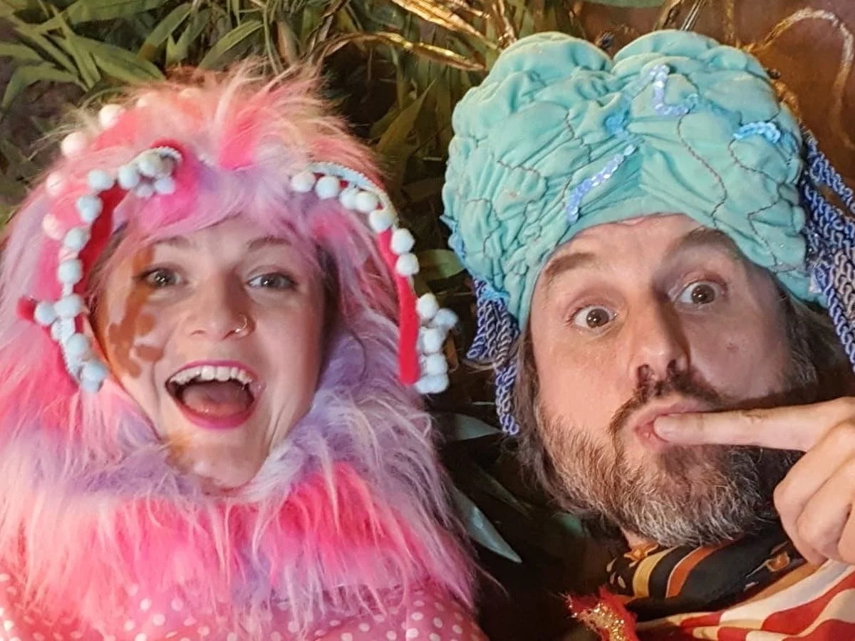Two people in colorful costumes taking a selfie. The person on the left has pink and purple fluffy hair with white pom-pom decorations, a painted face, and is smiling widely. The person on the right has a large turquoise headwrap with gold details, a
