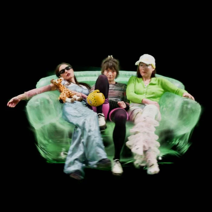 Three women sitting on a green velvet couch with a black background. The woman on the left is wearing sunglasses and holding a stuffed animal, the woman in the middle has a casual outfit with a headband, and the woman on the right is wearing a bucket hat and puffy boots. They appear relaxed and playful.