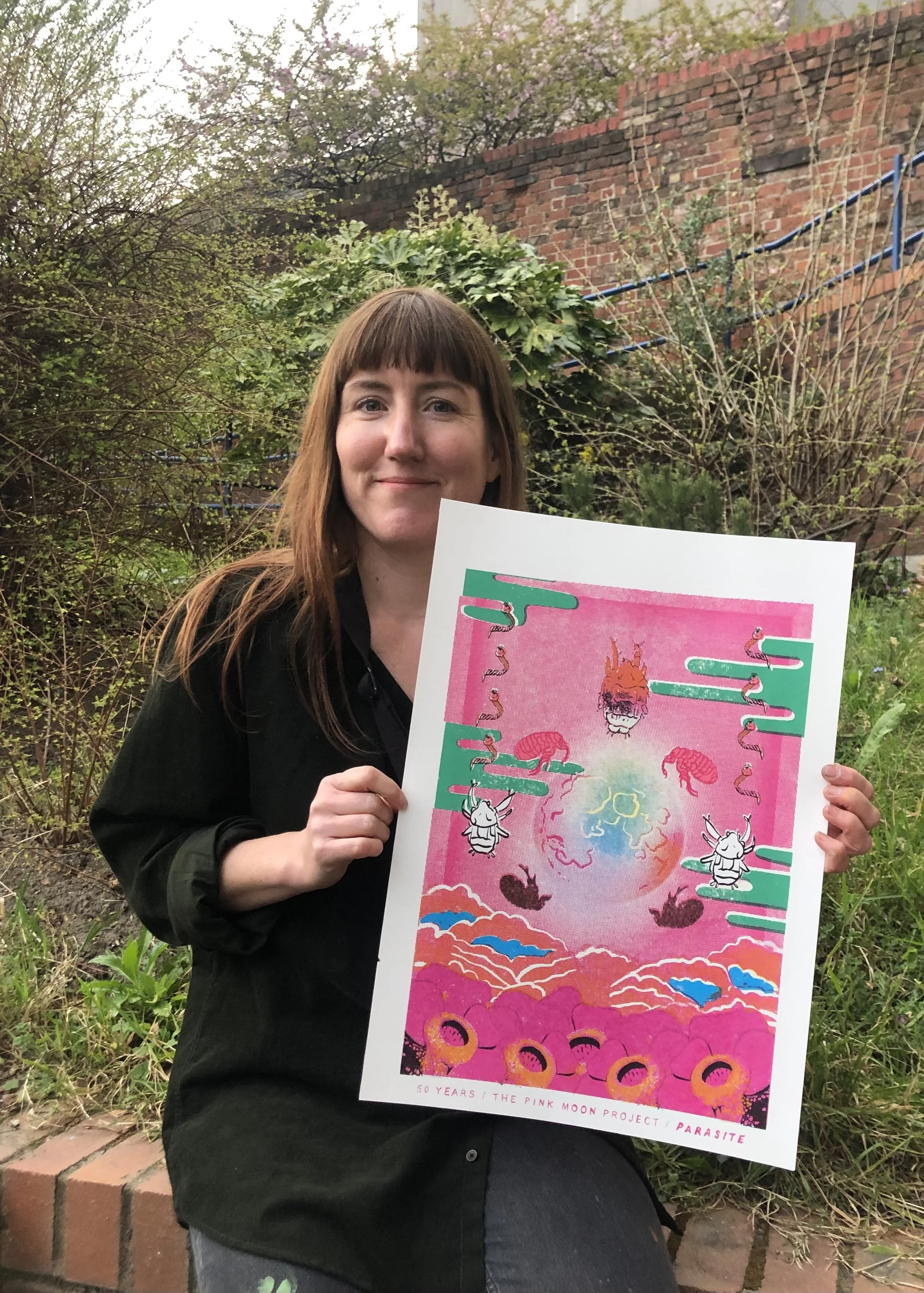 A woman with long brown hair holding a colorful poster outdoors, with trees and a brick wall in the background.