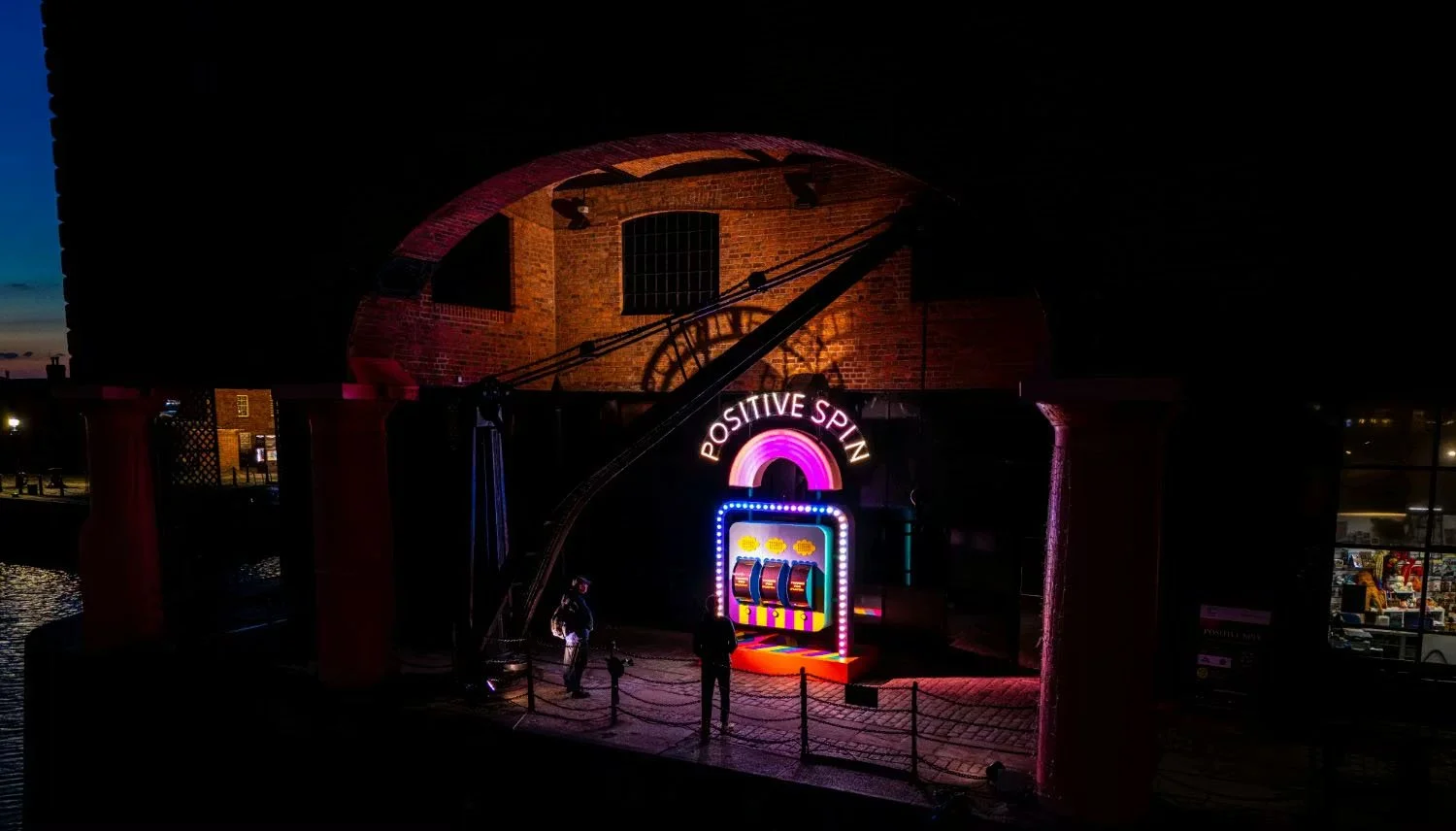 Night scene of a stage with neon signs reading 'Positive Spin' and a colorful slot machine display, with people standing nearby under a brick archway.