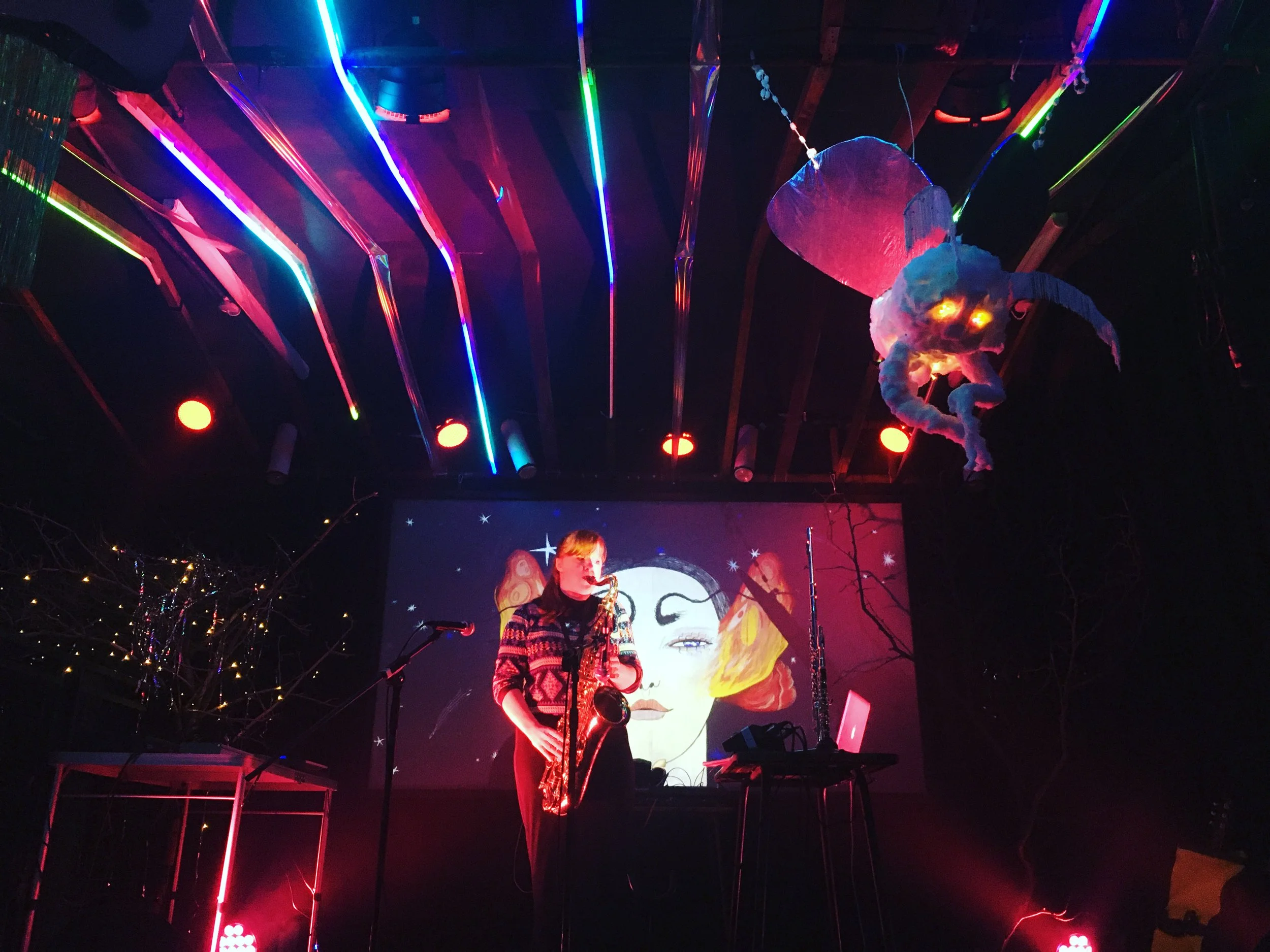 A musician playing a saxophone on stage with a colorful digital art projection of a woman's face behind them, neon lights hanging from the ceiling, and an animated jellyfish hanging from the ceiling.