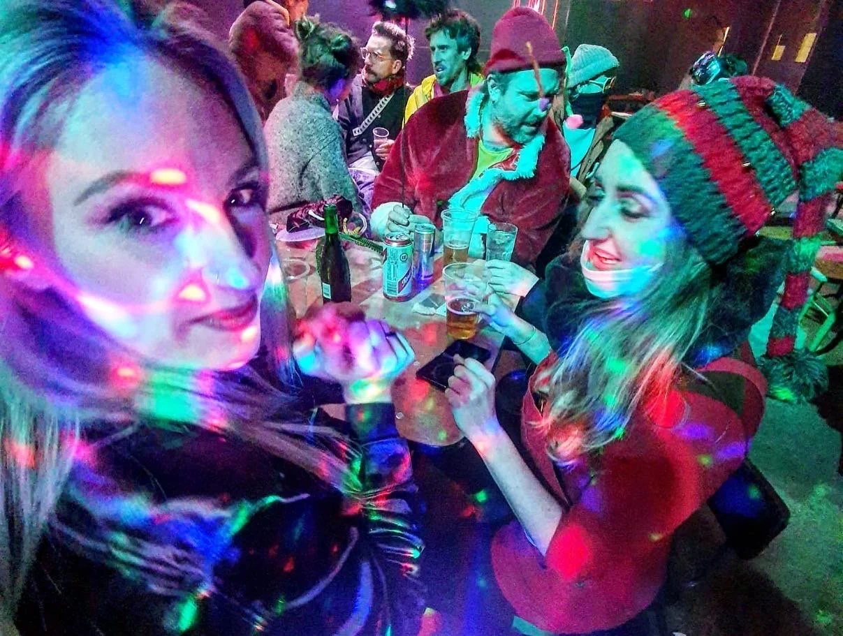 Two women at a party, one taking a selfie and the other looking at her phone, with colorful party lights and drinks on the table, one in a red Christmas sweater and hat, the other with blonde hair.