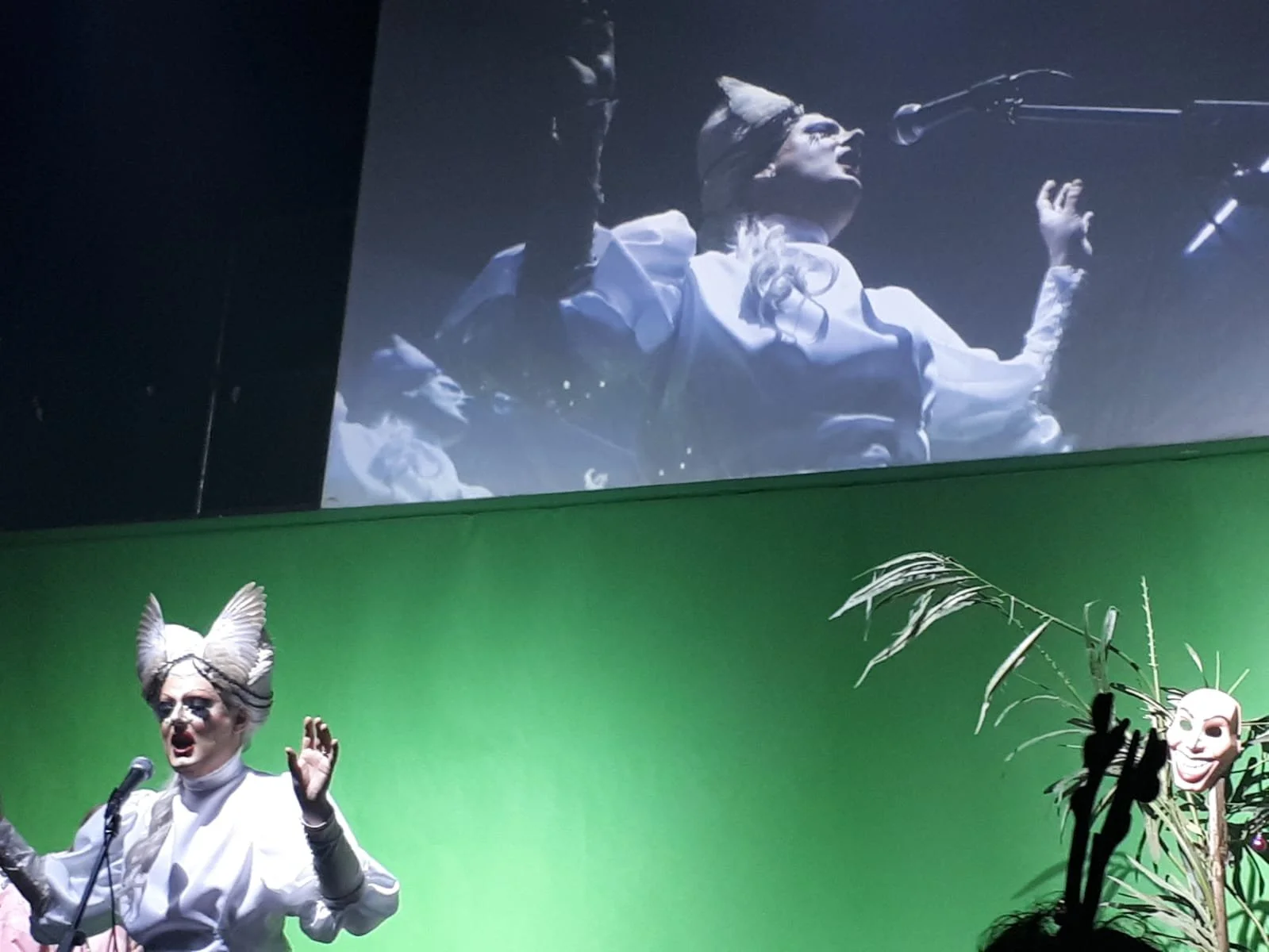 Performer dressed in a white costume with big feathers on their head speaking into a microphone on stage with a green backdrop and plant decorations, accompanied by a mask on a stick.
