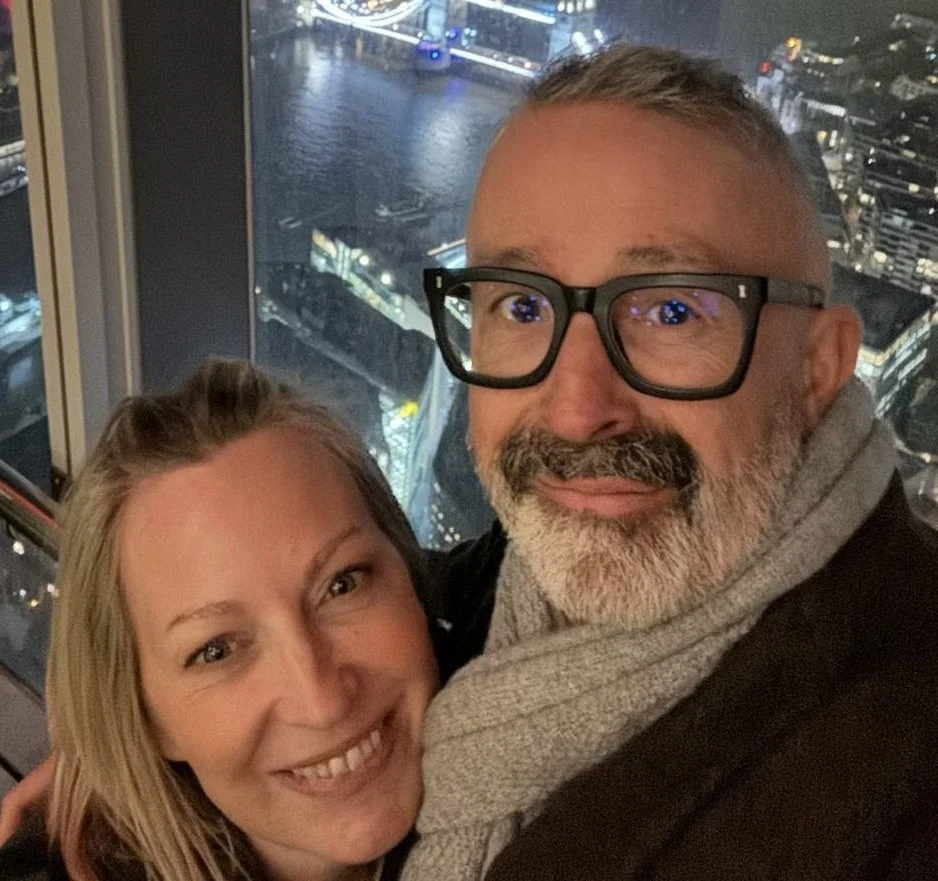 A smiling woman and a smiling man with glasses taking a selfie in front of a cityscape at night from a high vantage point.