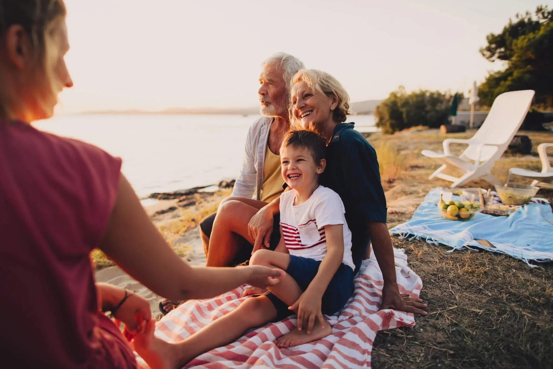 A family picnic on a beach during sunset, with elderly couple, young boy, and woman sitting on a striped blanket, holding hands, with food and chairs nearby.