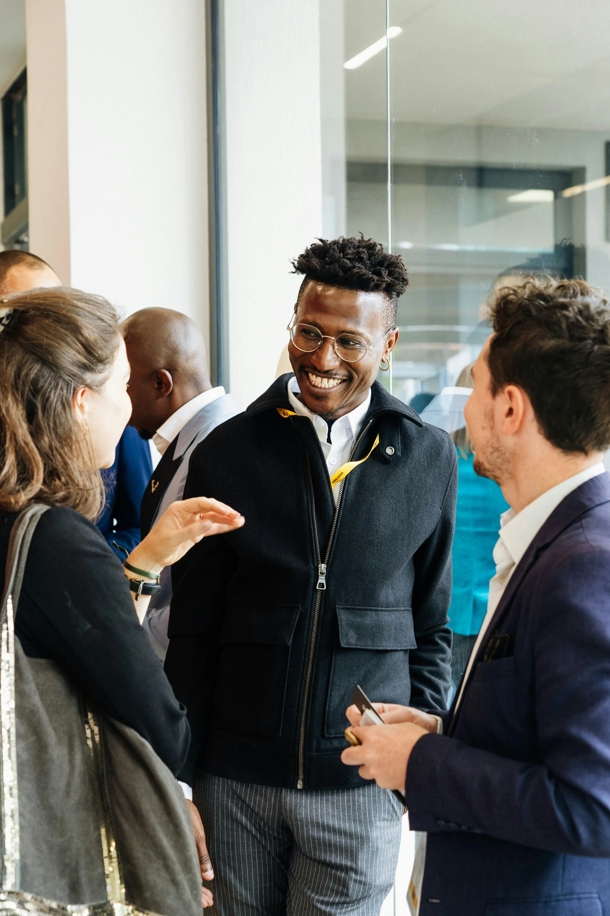 A group of diverse young professionals, including a smiling Black man with glasses, are engaged in conversation at a networking event or conference.