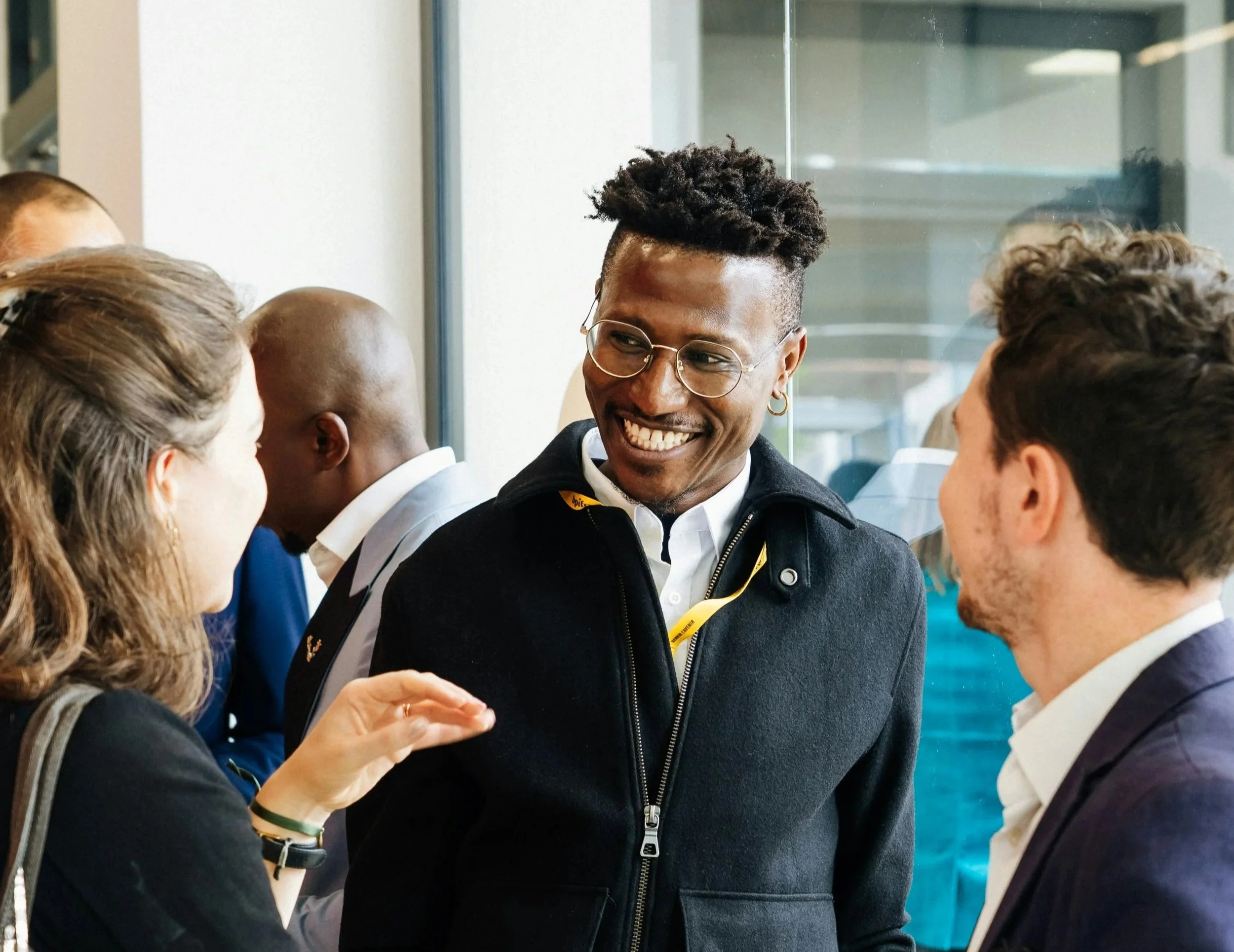 A group of diverse people engaged in conversation indoors, with a smiling person at the center.
