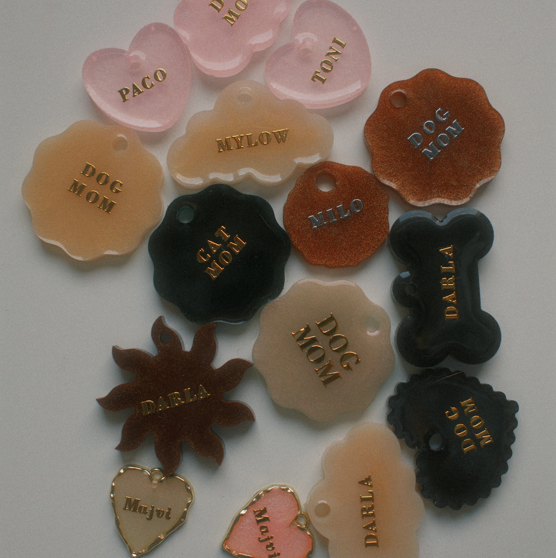 Verschiedene Knochen- und Hundeanhänger aus transparentem und opakem Material in verschiedenen Farben mit goldener oder silberner Beschriftung. Einige Anhänger sind mit Namen wie 'PACO', 'TOBY', 'MYLO', 'DARCY' und 'MAJOR' beschriftet.