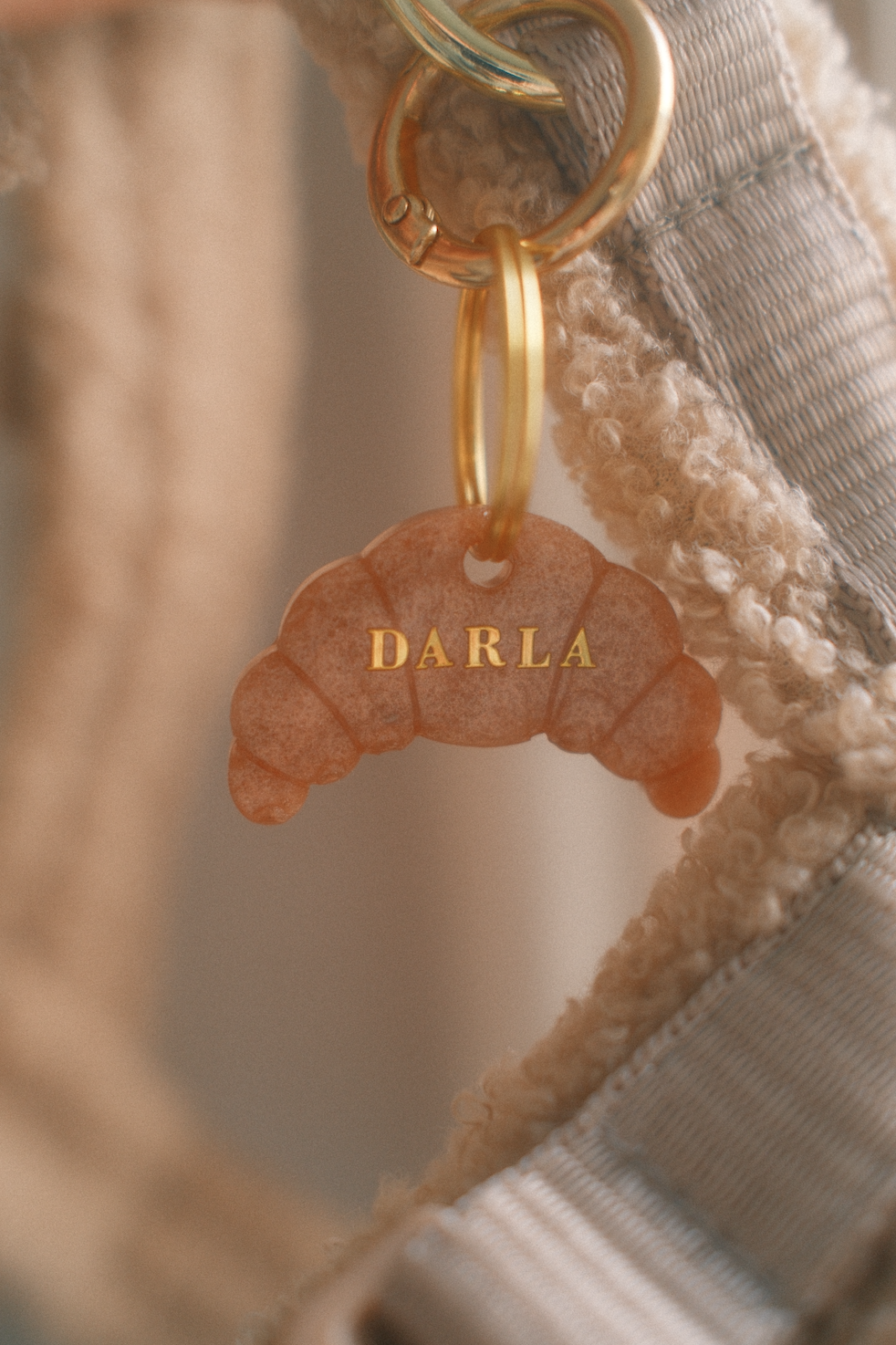 Ein Schlüsselanhänger in Form eines Croissants mit dem Namen 'DARLA' in goldener Schrift, befestigt an einem Schlüsselring.