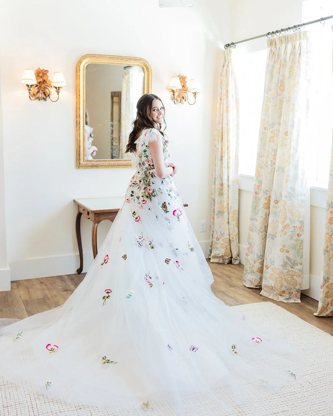 A young woman in a white, floral embroidered dress standing in a bright room with cream-colored curtains, a mirror, and wall lamps.