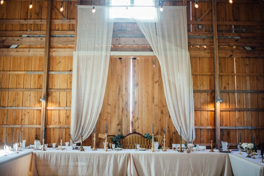 Long banquet table set for a formal event inside a rustic barn with wooden walls and a high ceiling, decorated with candles, white tablecloths, and floral arrangements. A velvet tufted sofa is positioned at the head of the table, framed by large white curtains.
