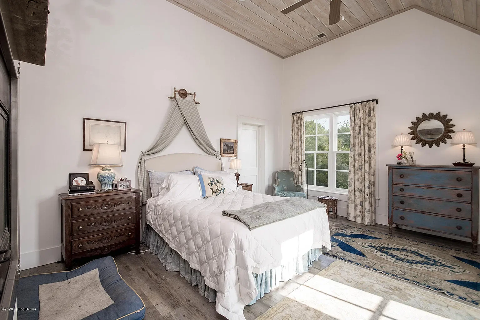 A bedroom with a large bed that has a white quilt and decorative pillows, two nightstands with lamps, an antique dresser, a blue armchair near a window, patterned curtains, and a wooden ceiling.