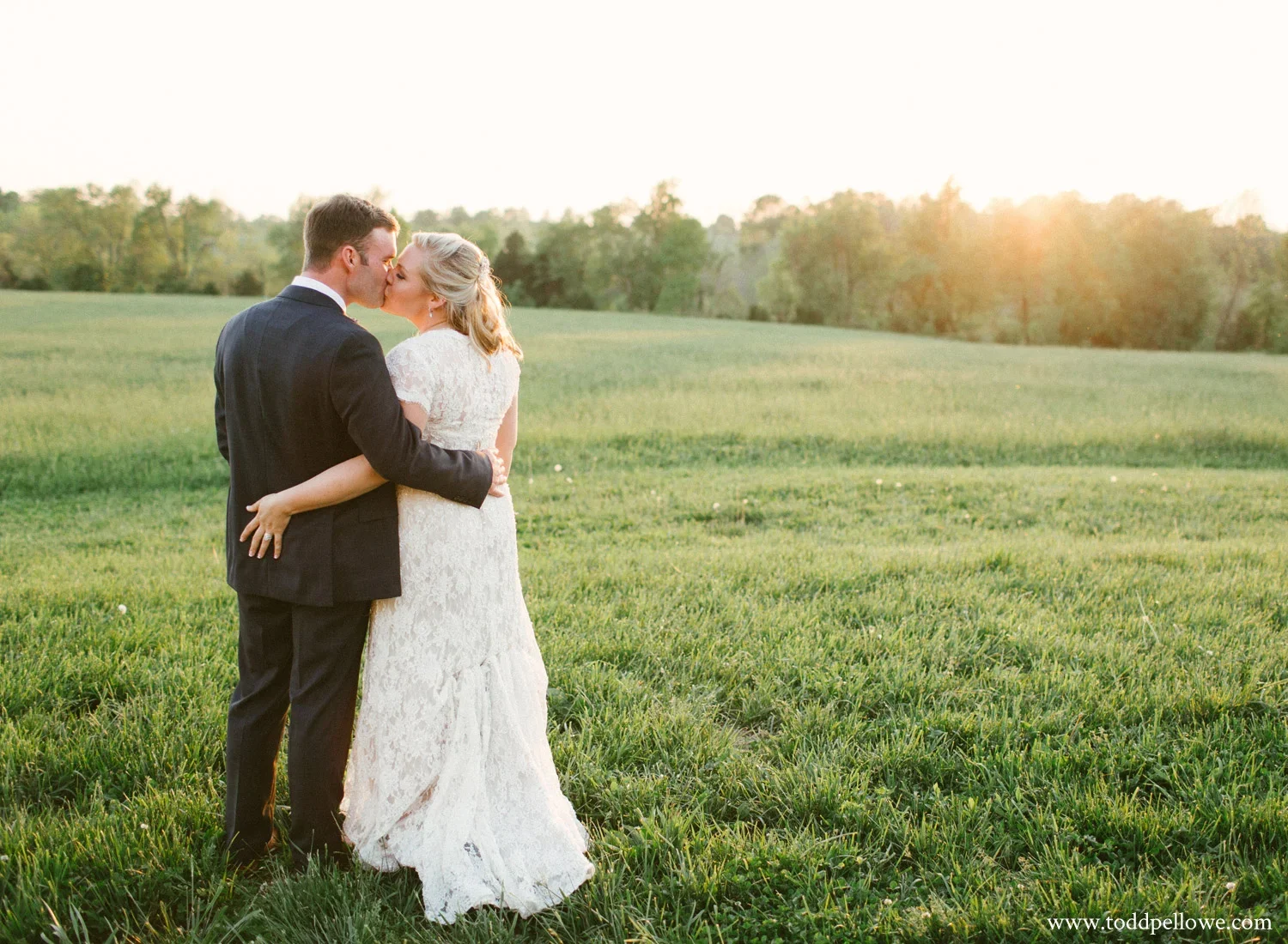 A newlywed couple sharing a kiss in a green field during sunset, the bride in a white lace wedding dress and the groom in a dark suit.