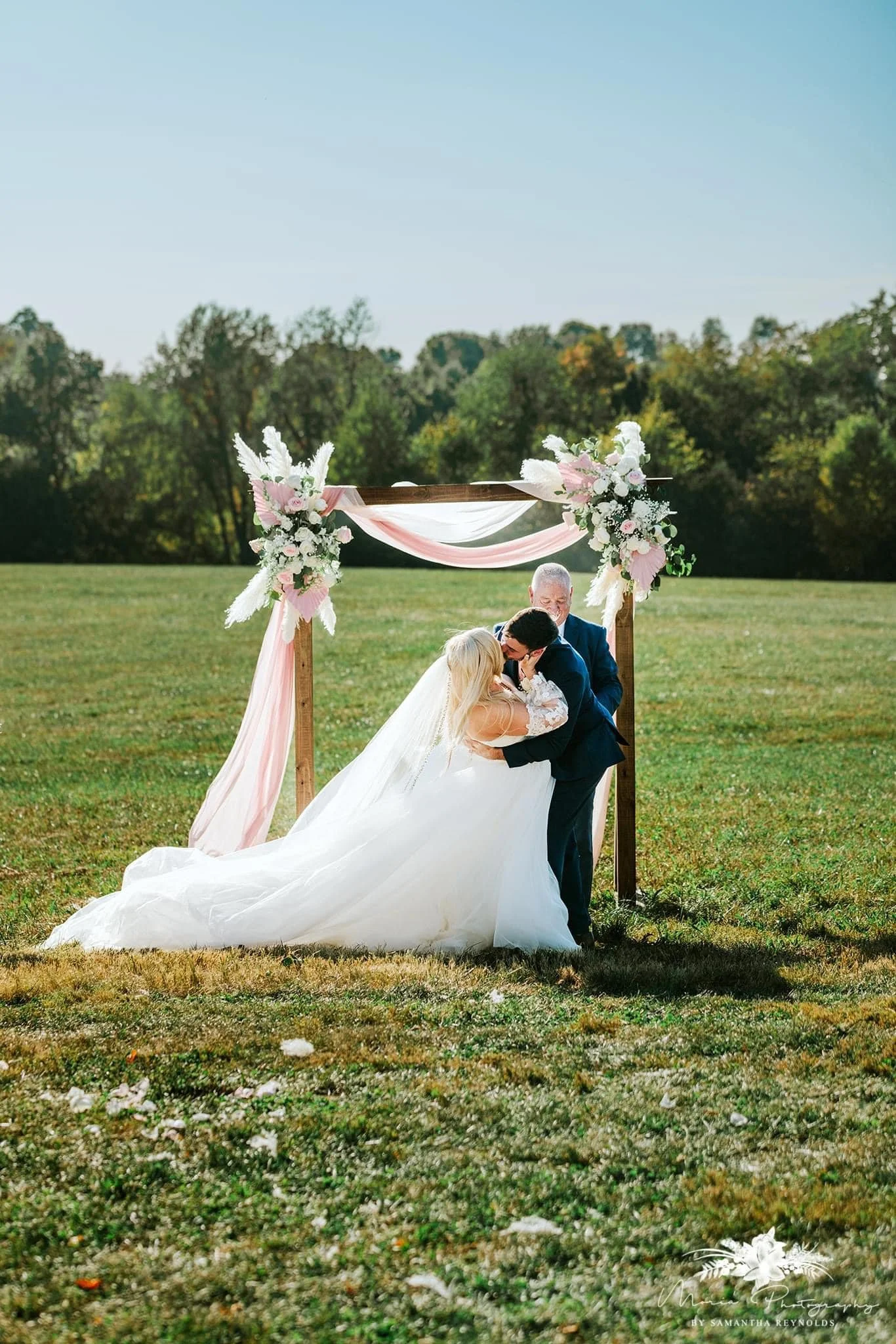 A wedding couple kissing under a decorated wooden arch outdoors on a grassy field with trees in the background.
