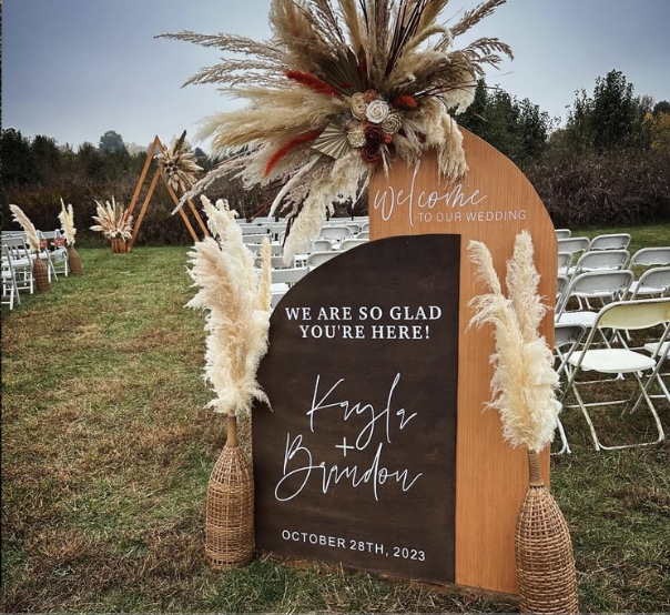 Wedding welcome sign with dried pampas grass and floral arrangements in woven vases, set outdoors on grass with chairs and an arch in the background.