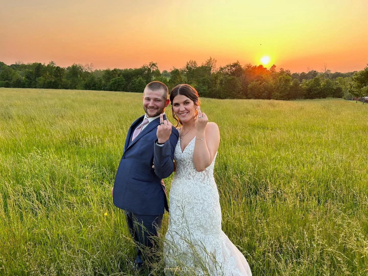 A newlywed couple standing in a grassy field at sunset, both showing their middle fingers, smiling.