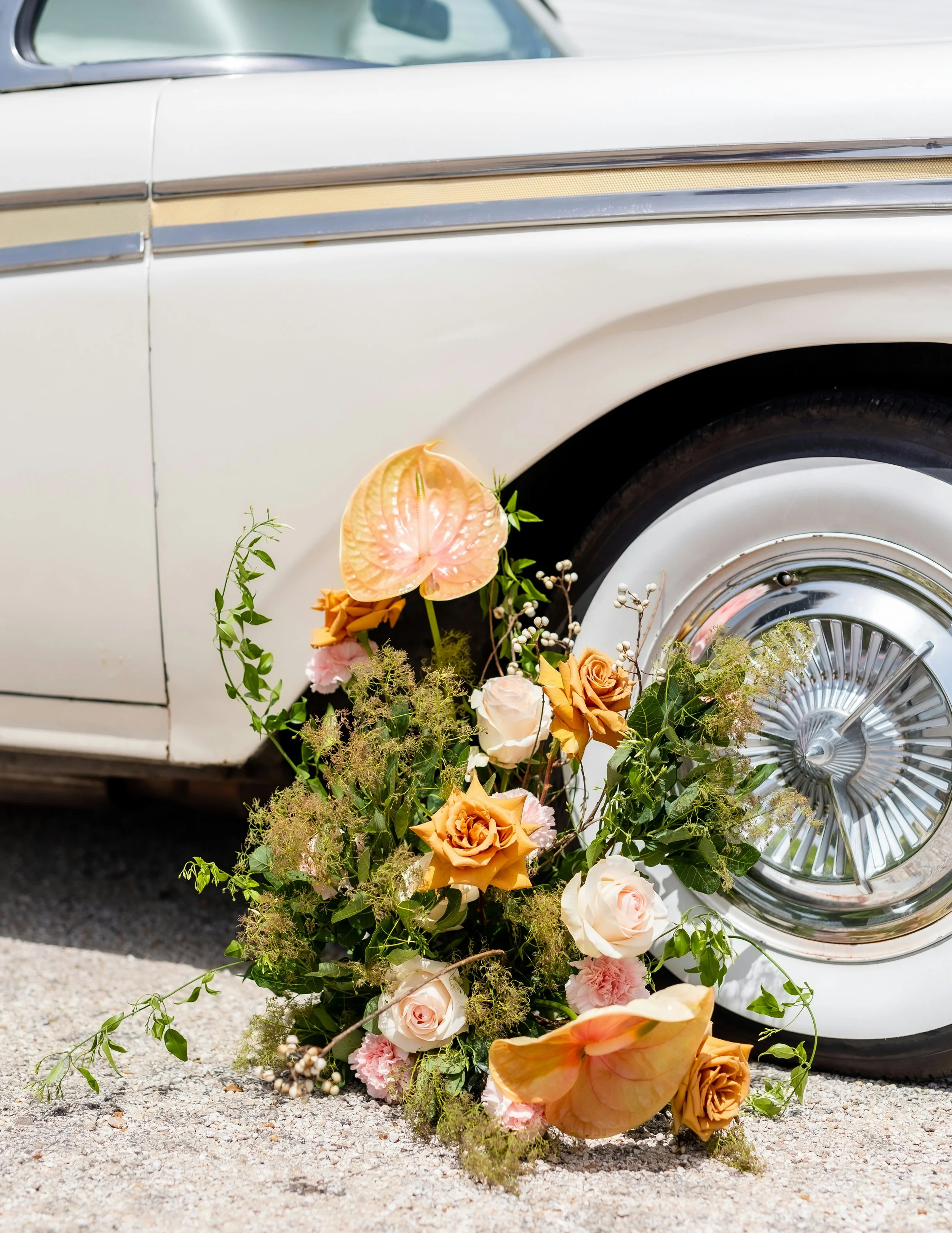 A floral arrangement of roses and other flowers lies on the ground near a vintage white car with a decorated wheel, possibly part of a wedding or special event.