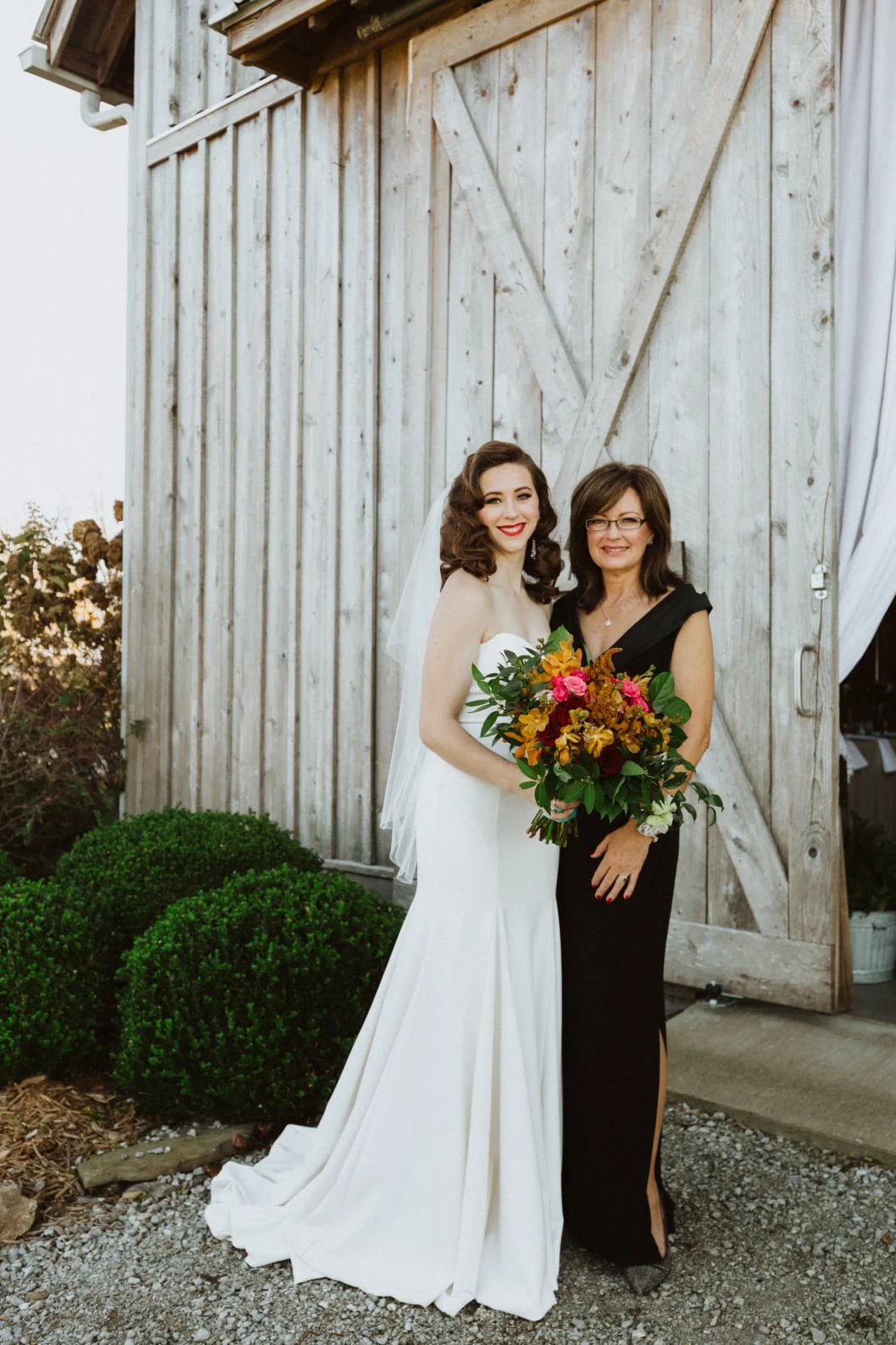 A bride with dark brown wavy hair and a white wedding gown stands next to a woman with glasses and a black dress, holding a bouquet of flowers, in front of a rustic wooden barn door.