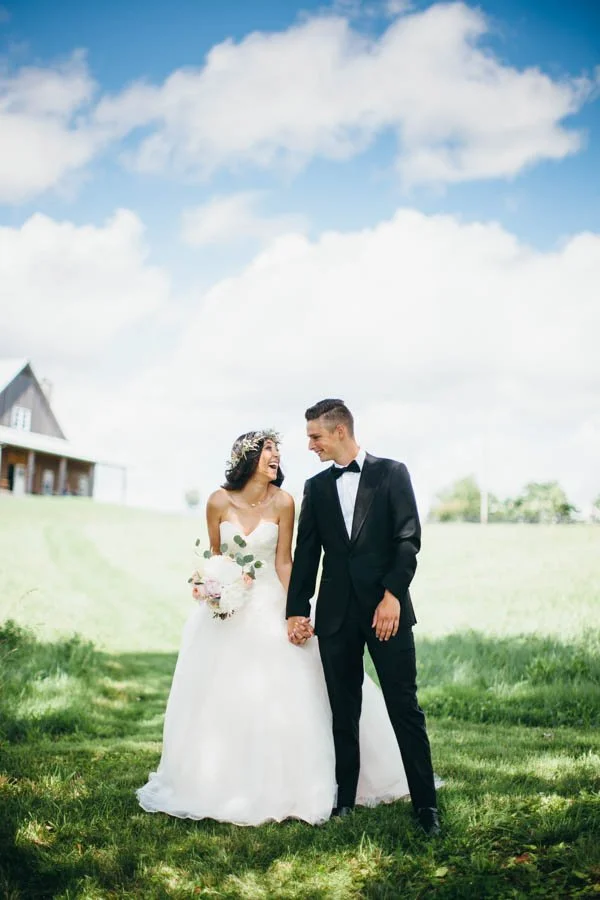A bride and groom holding hands outdoors on a sunny day with a house and green landscape in the background.