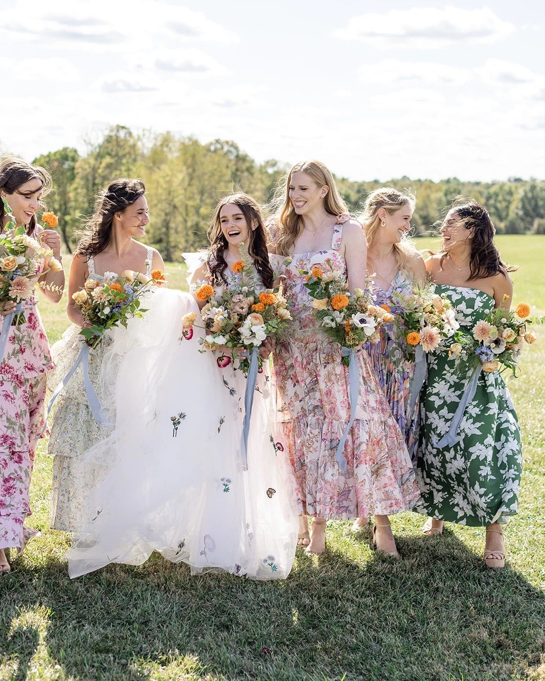 A group of six women, including a bride in a white wedding gown, walking outdoors on a sunny day. They are all holding colorful floral bouquets and are dressed in floral-patterned dresses, smiling and enjoying each other's company in a grassy field with trees in the background.