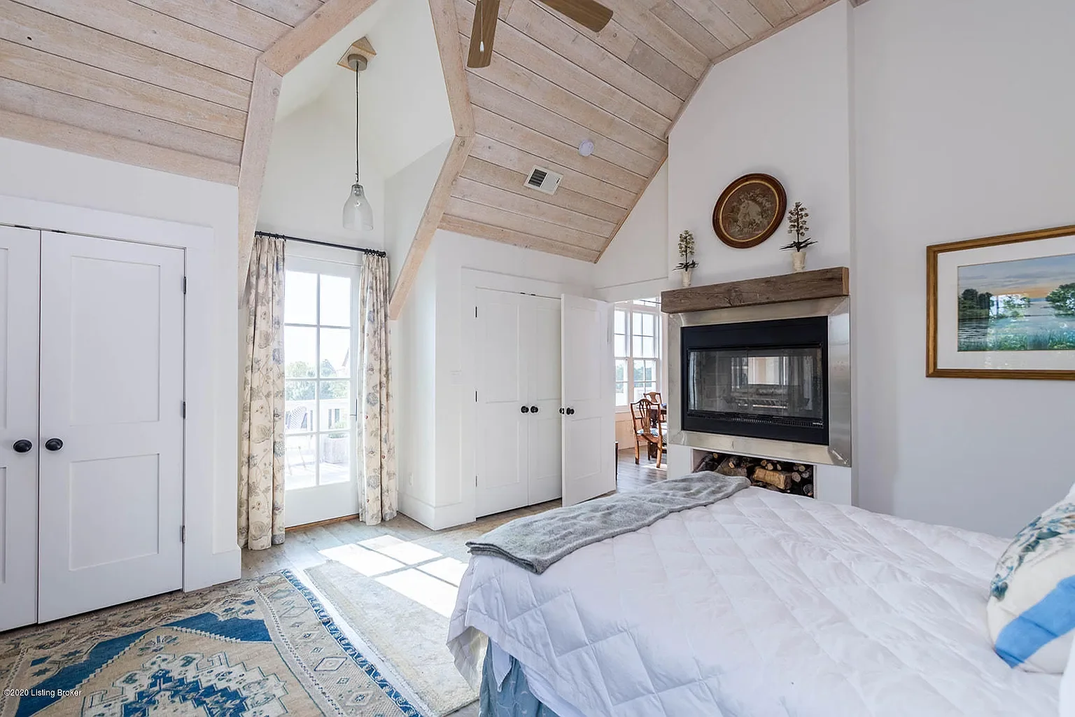 A cozy bedroom with a vaulted wood ceiling, white walls, a fireplace with wood storage underneath, a bed with white bedding and a gray throw, a window with floral curtains, and a door leading outside.