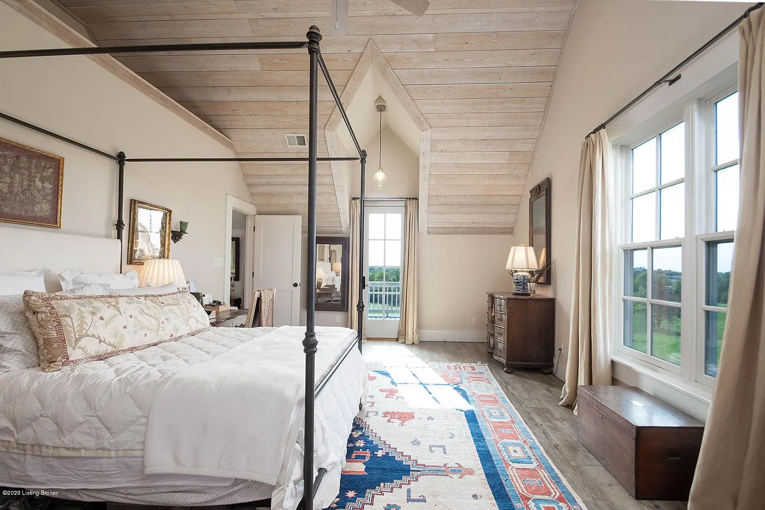 A bedroom with a white bed, beige and floral pillows, wooden flooring, a rug with geometric patterns, large window with cream curtains, a dresser with a lamp, and a balcony door with vision of outdoors.