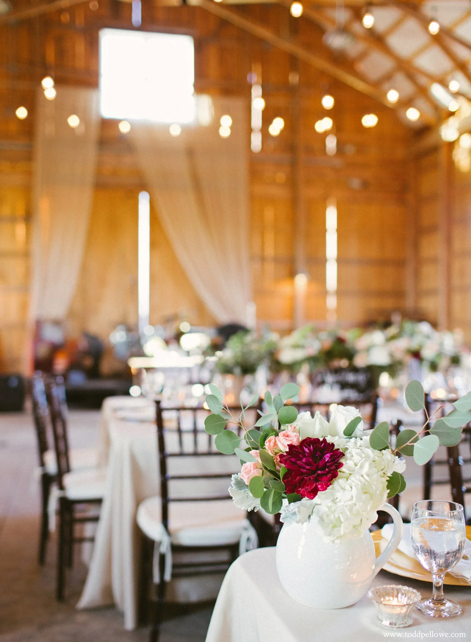 A floral centerpiece with deep red, pale pink, and white flowers, along with eucalyptus leaves, on a table at a rustic wedding reception with wooden walls and high ceiling.