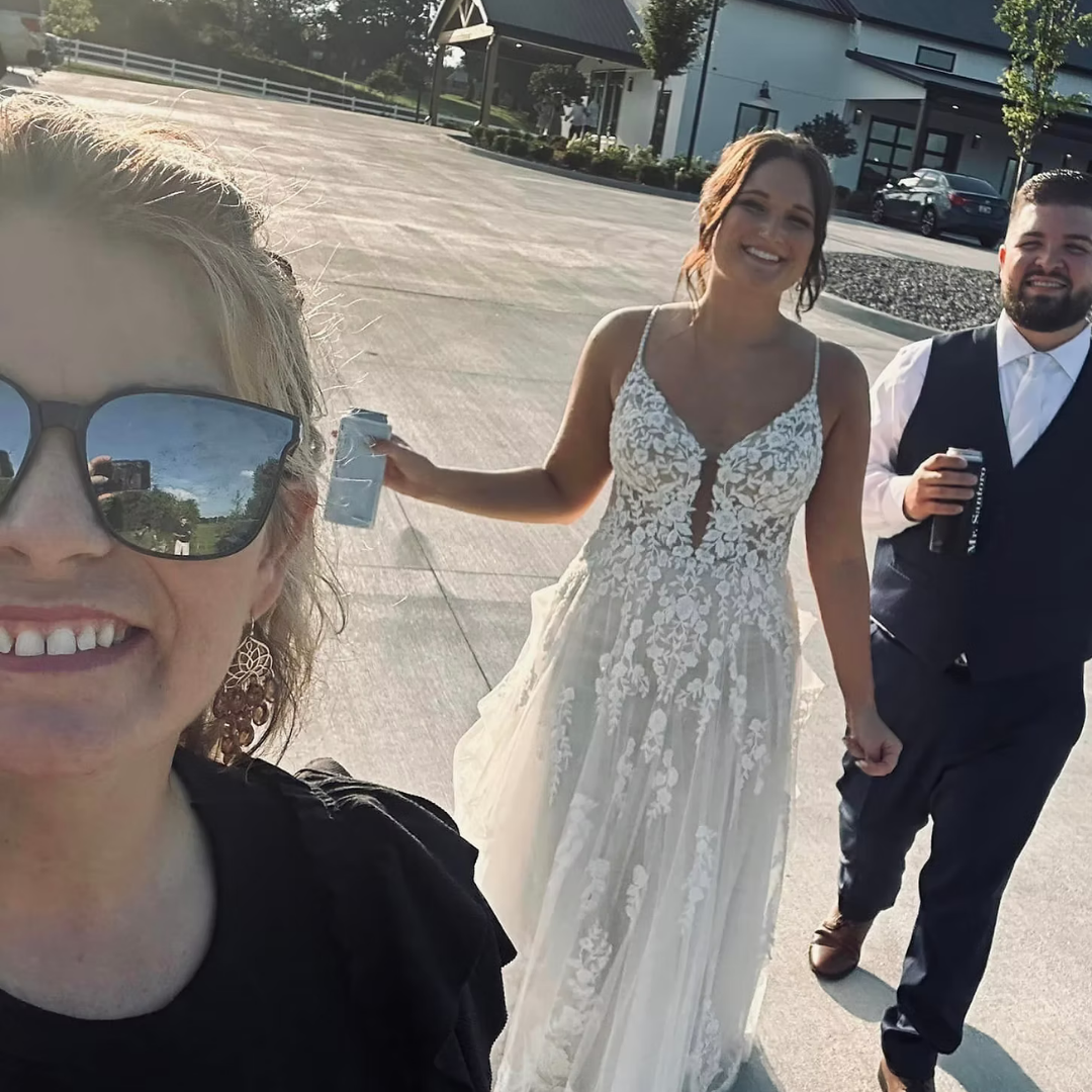 Three people taking a selfie outdoors, with a smiling woman in a wedding dress, a man in a white shirt and vest holding a drink, and a woman wearing sunglasses and earrings.