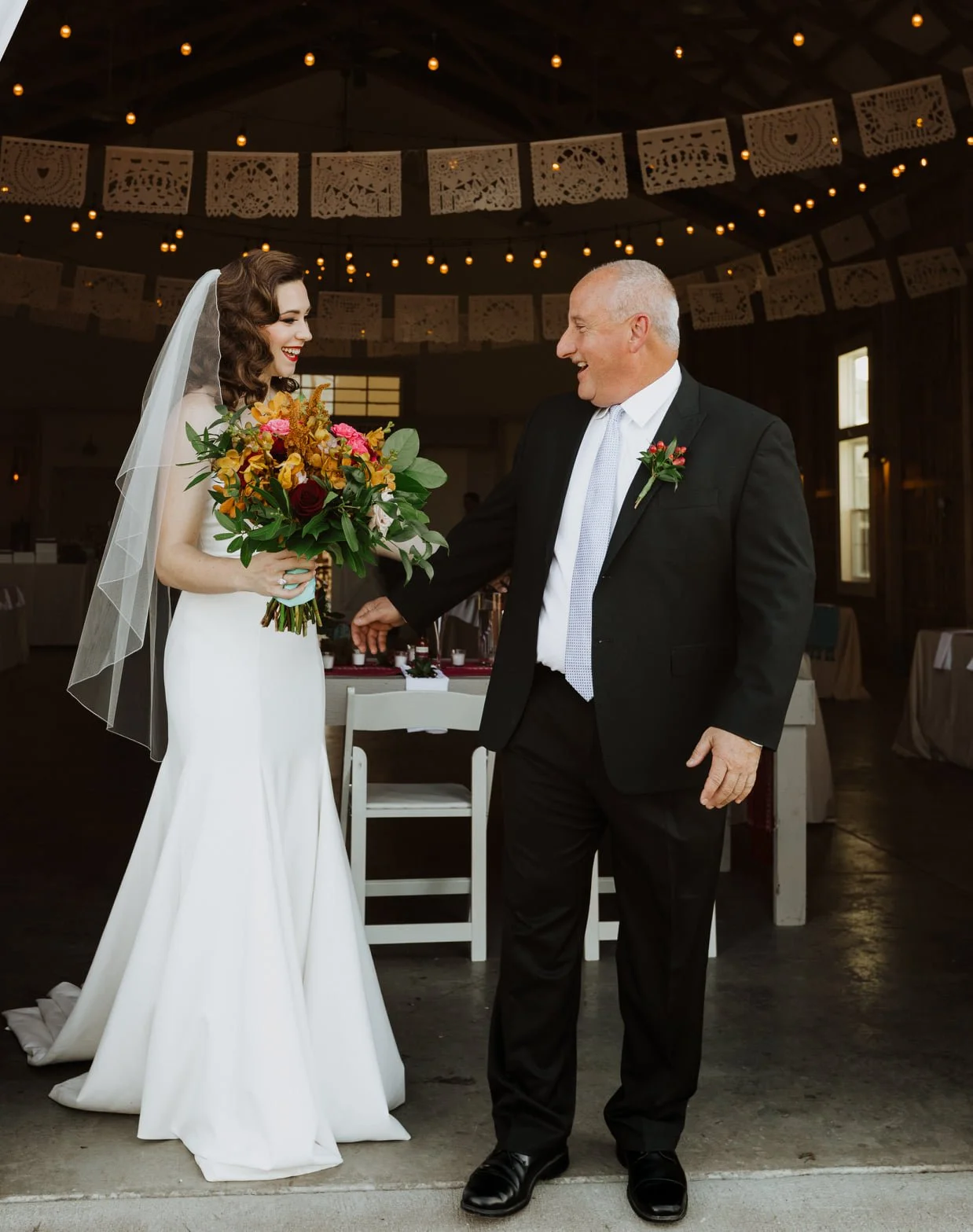 A bride in a white wedding dress holding a bouquet of colorful flowers, smiling at an older man in a black suit with a white shirt and tie, inside a decorated venue with hanging paper banners and string lights.