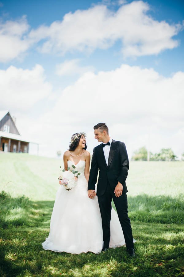 A bride and groom holding hands, smiling at each other outdoors on a sunny day, with a house and trees in the background.