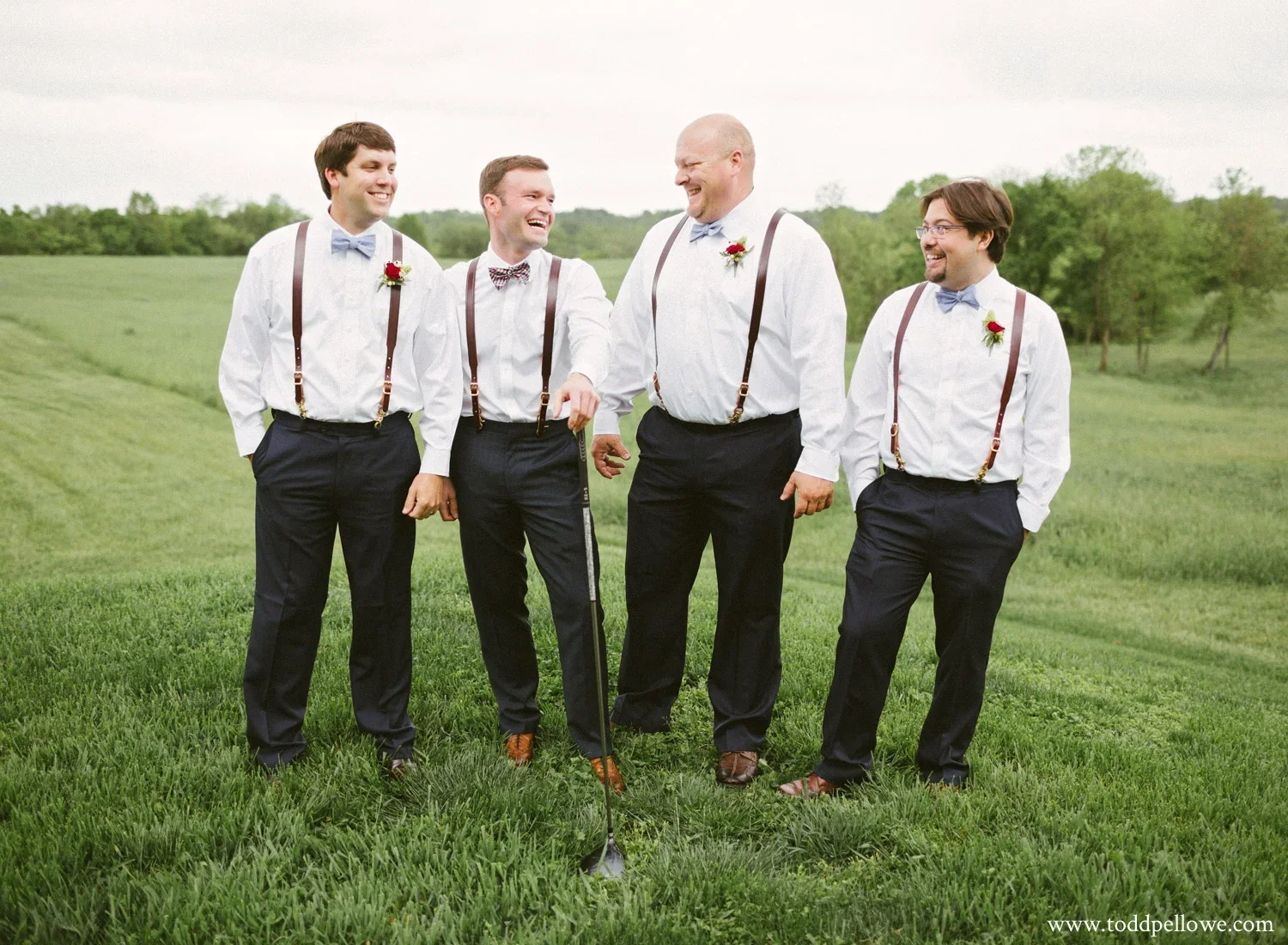Group of five men dressed in white shirts, suspenders, and bow ties standing on a grassy field, smiling and talking, with trees and overcast sky in the background.