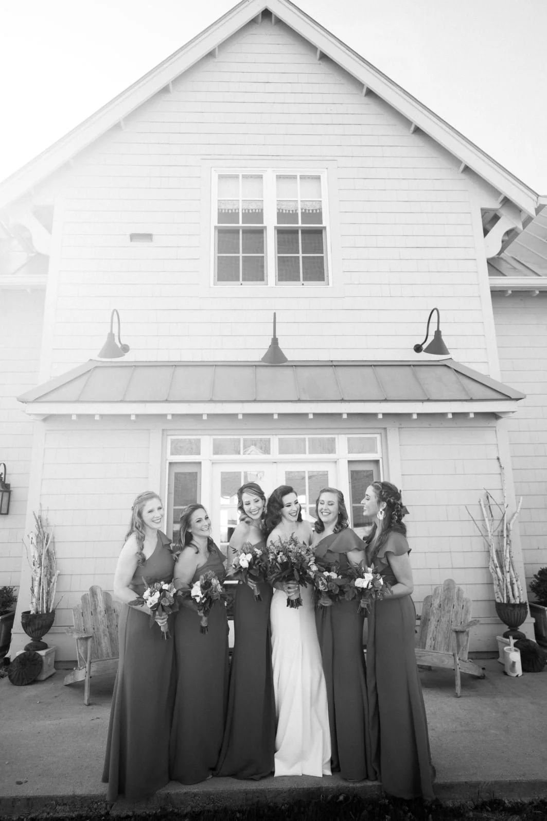 A wedding group photo of six women, with one bride in a white gown and five bridesmaids in long dresses, standing outside in front of a house with a sloped roof and large windows, all holding bouquets and smiling.