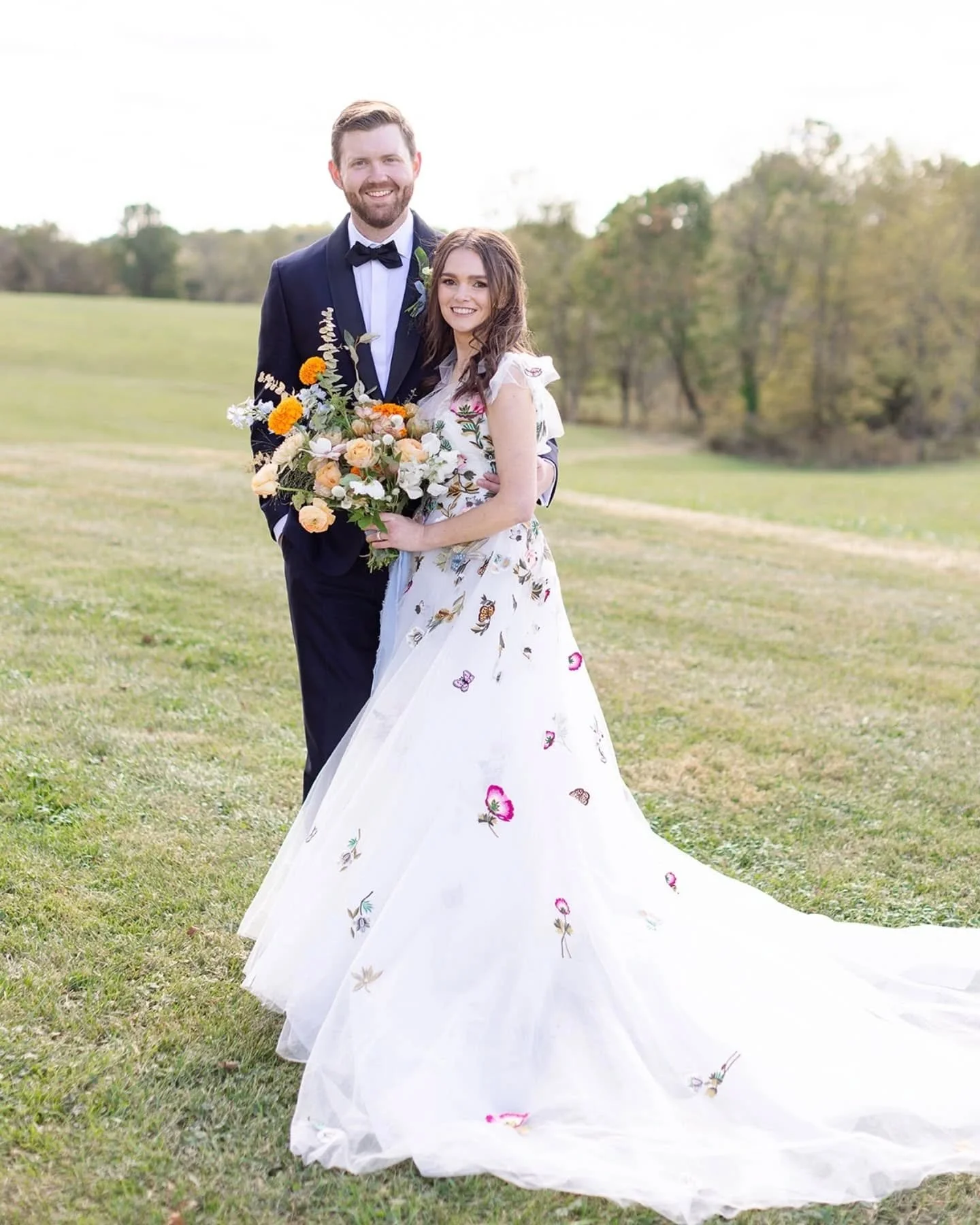 A bride and groom stand on a grassy field, smiling for a picture, with trees in the background. The bride wears a white wedding dress with colorful embroidery and a long train, holding a bouquet. The groom wears a black tuxedo with a bow tie.