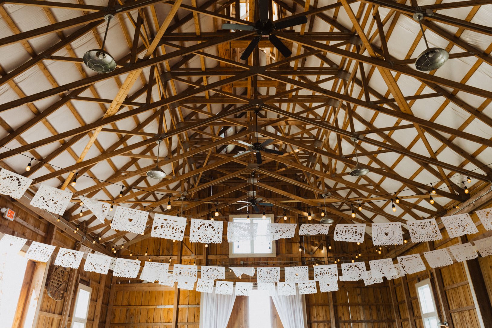 Decorative white paper banners with intricate cut-out designs hanging in a rustic barn with exposed wooden beams, string lights, and windows.