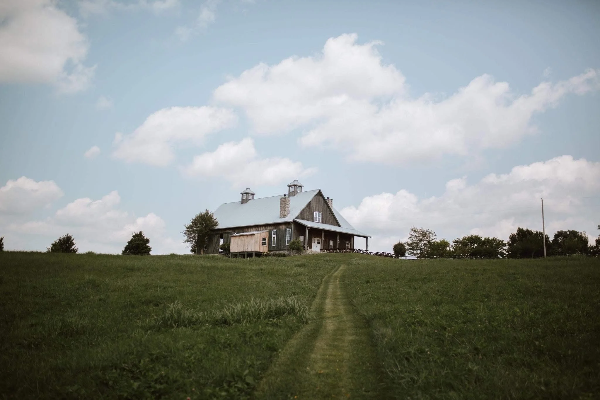 A wooden house on a grassy hill with a dirt path leading to it, under a partly cloudy sky.
